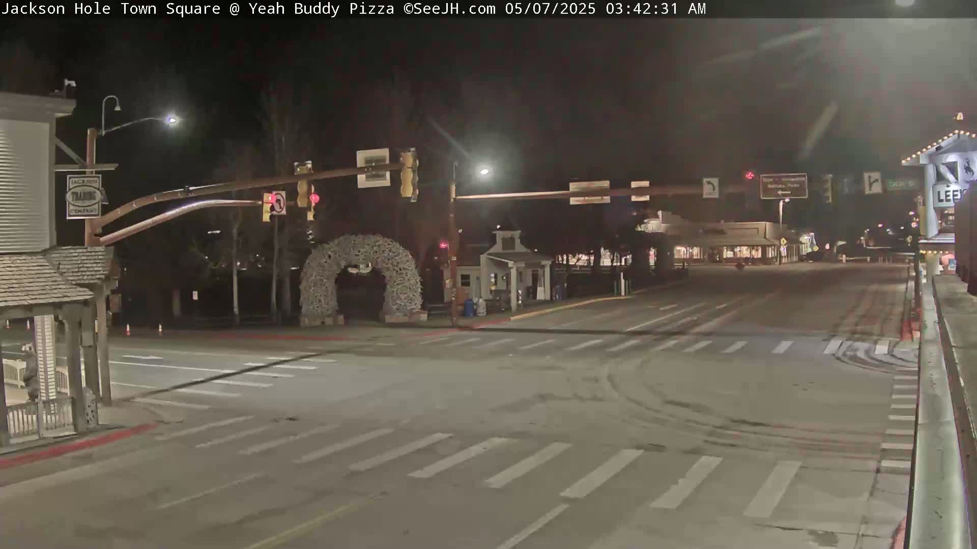 A nighttime street scene shows a mostly empty town square intersection with traffic lights and tire tracks in the snow.
