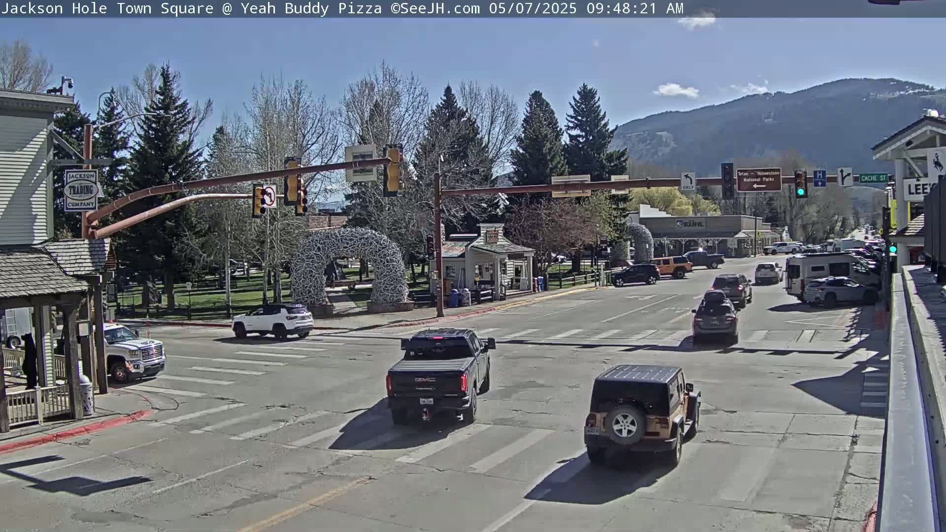 A sunny day reveals a town square intersection with several vehicles, a park-like area with an arched structure, and buildings in the background, including mountains in the distance.