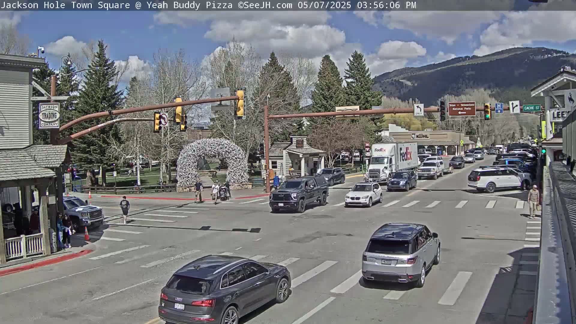 A partly cloudy afternoon scene shows a town square intersection with several vehicles, pedestrians, and buildings, with mountains visible in the background.