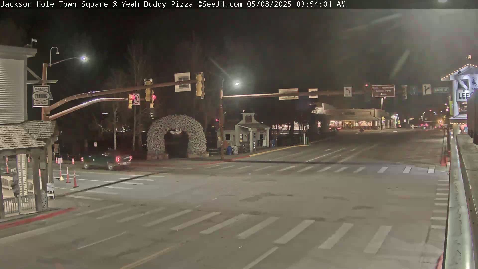 A nighttime view of a town square intersection with a few cars and traffic signals, under dark, clear skies.