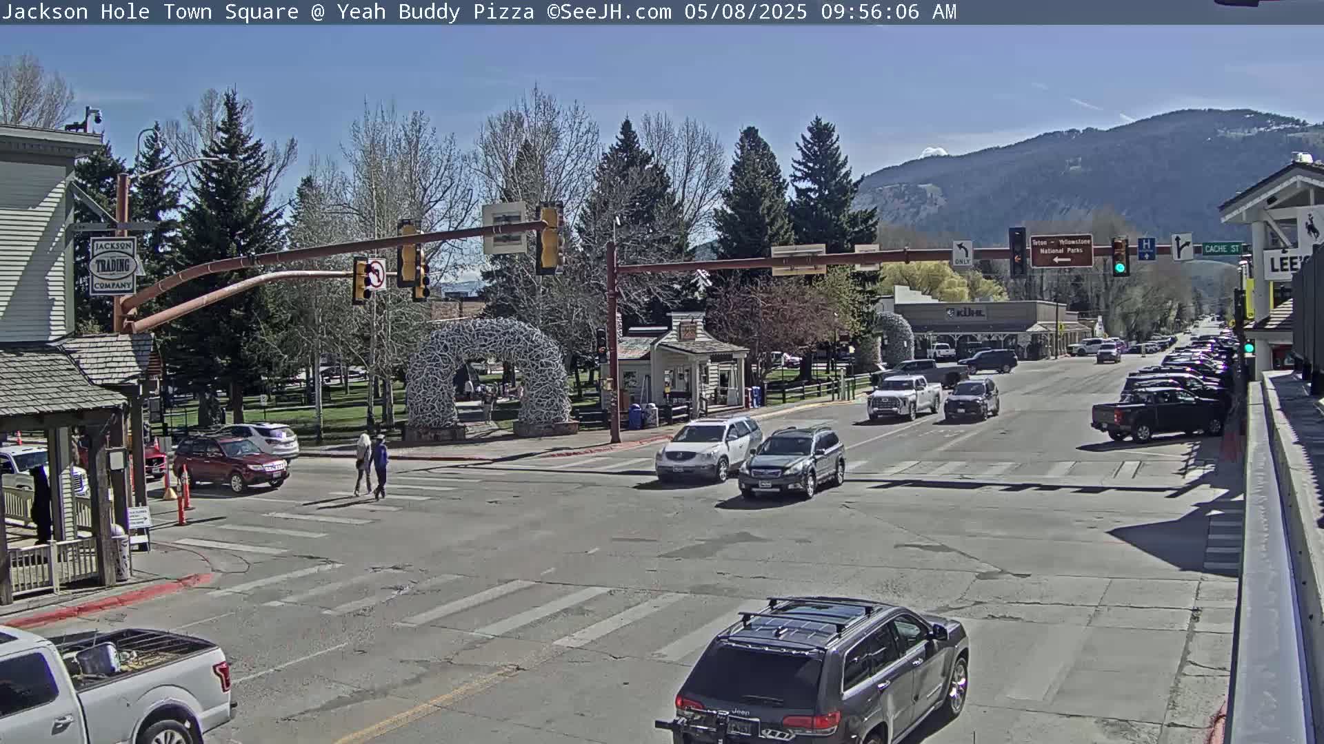 A sunny day in a town square shows several cars at a multi-way intersection with pedestrians crossing, and mountains in the background.