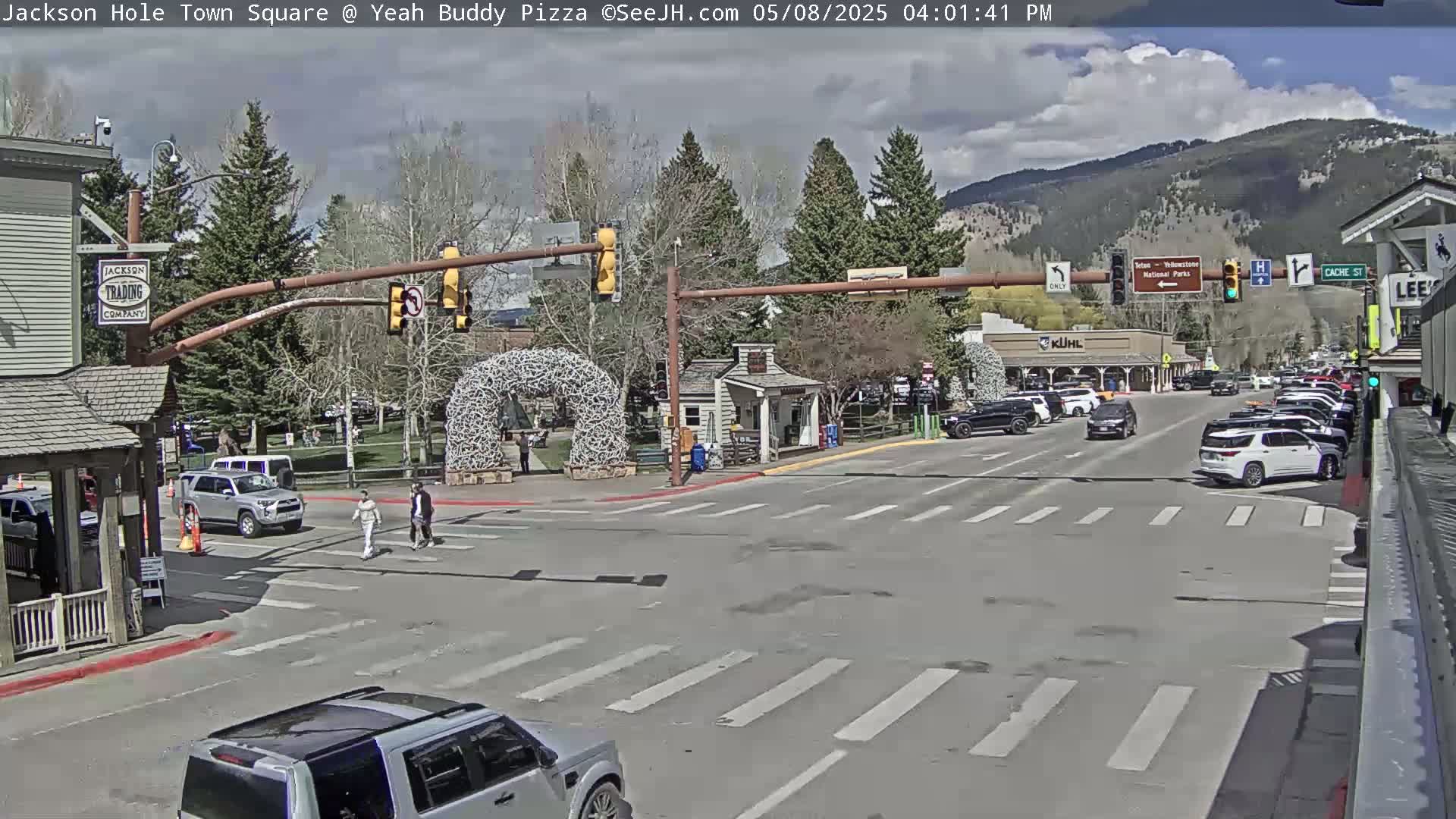 A partly cloudy day reveals a town square intersection with several vehicles, pedestrians, and buildings, including a small park with a large arch.