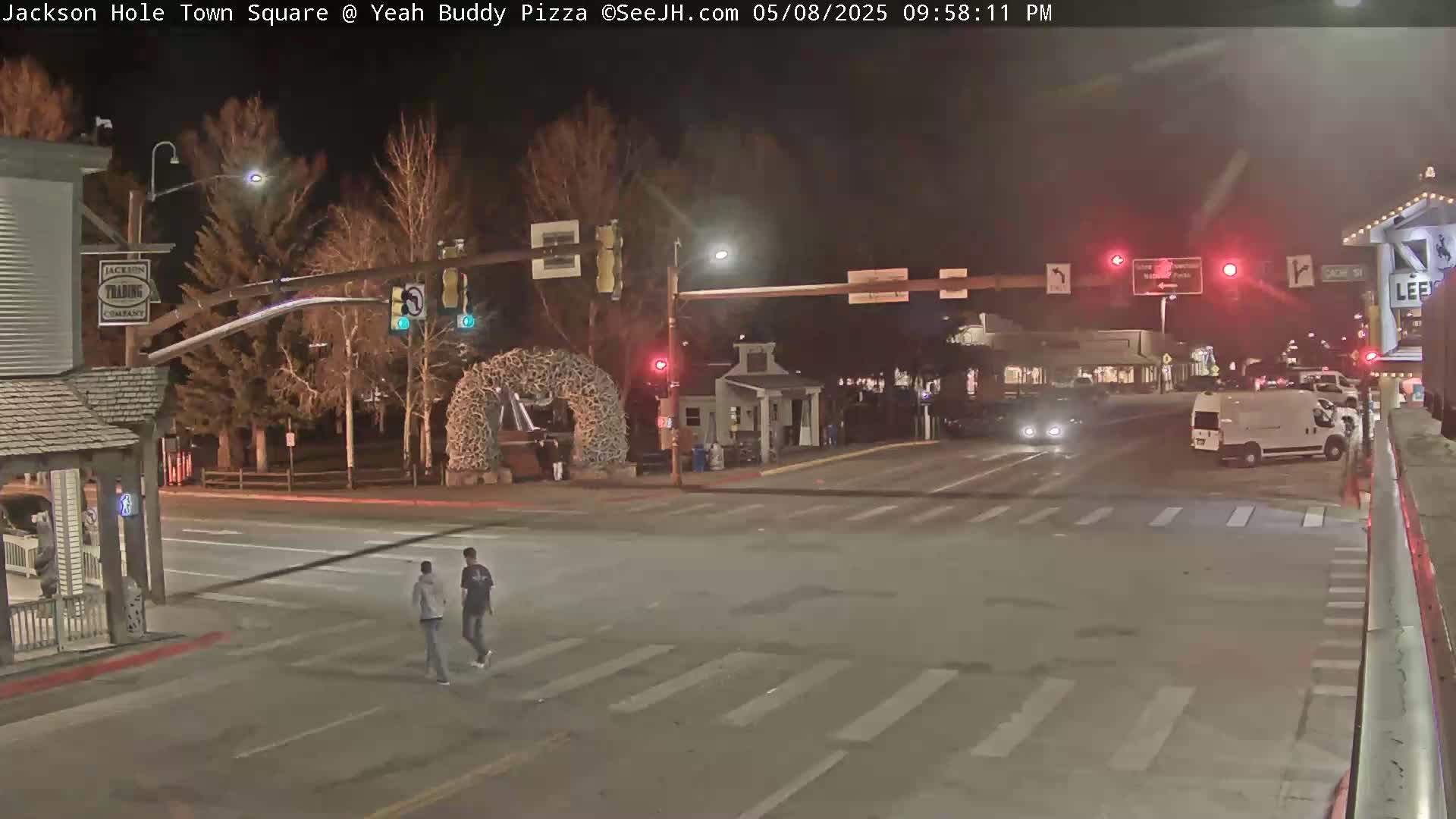 At night, two pedestrians cross a street intersection in a town square, with a few vehicles visible and bare trees in the background.