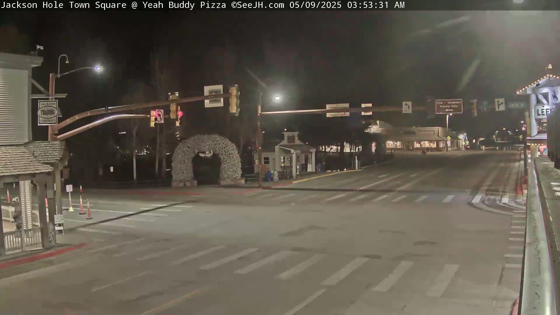 A nighttime, empty town square intersection features a traffic light and an antler archway under streetlights.