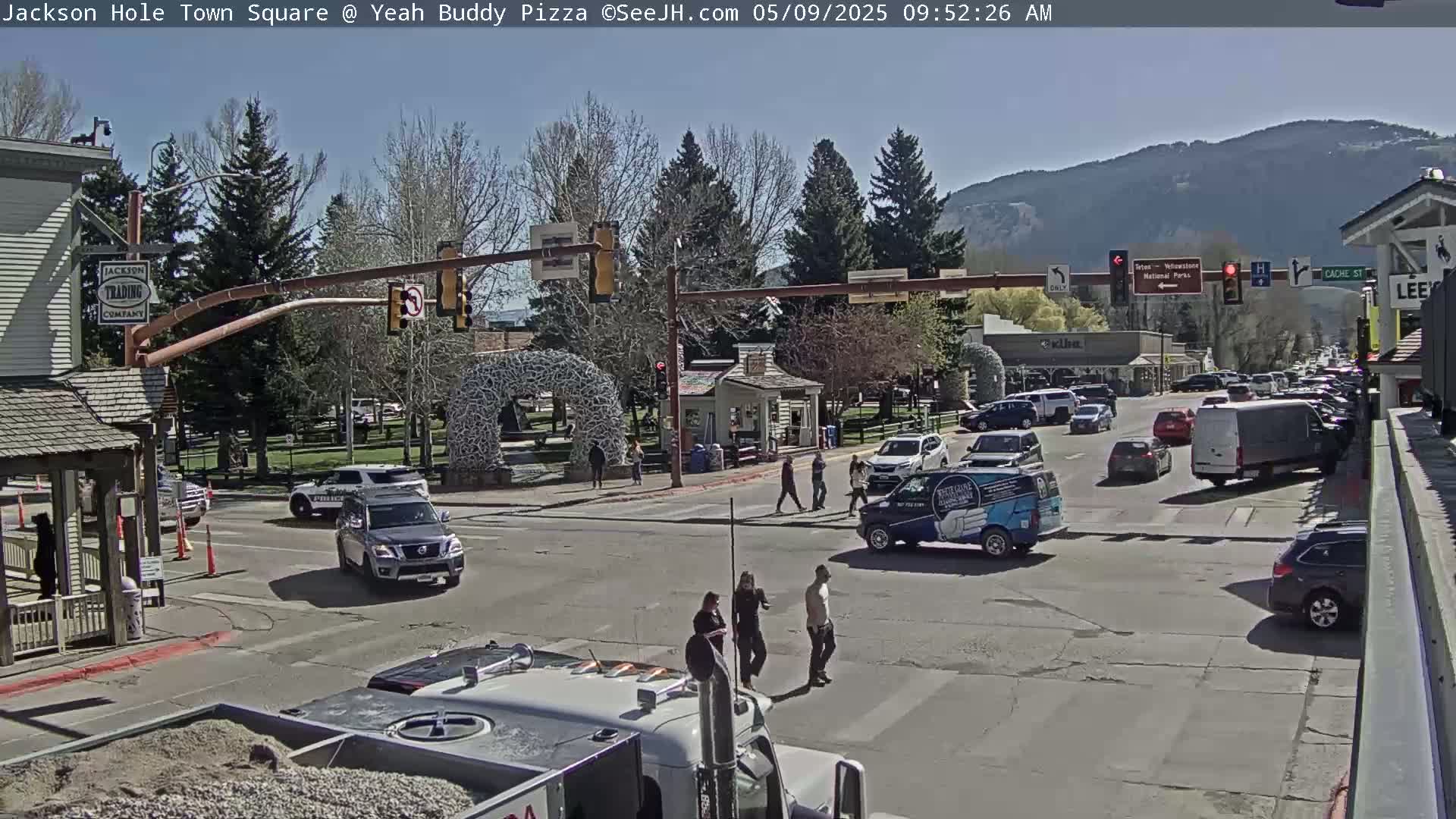 A sunny town square scene shows pedestrians crossing a street intersection with several vehicles, including police cars and vans, under a clear sky and mountains in the background.