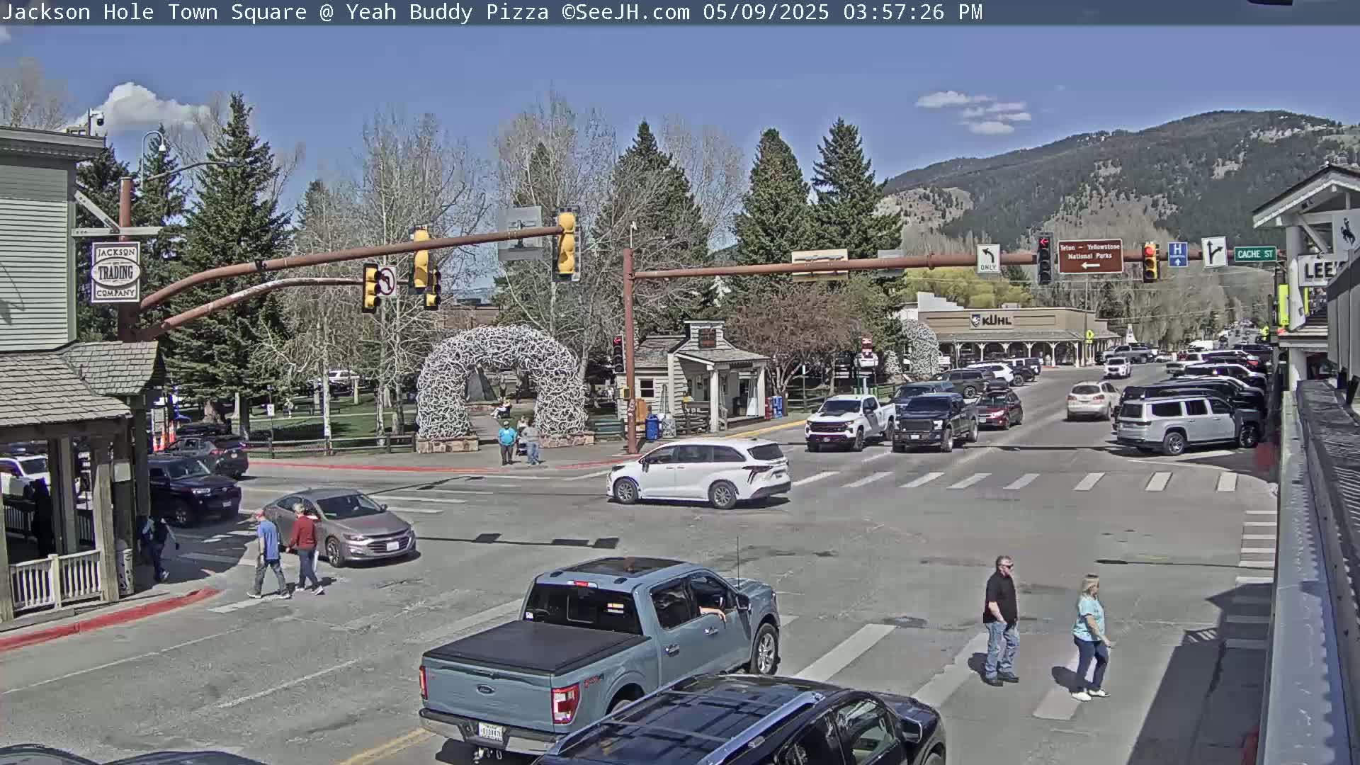 A sunny town square scene shows several vehicles at a four-way intersection, with pedestrians crossing the street and some parked cars lining the edges.