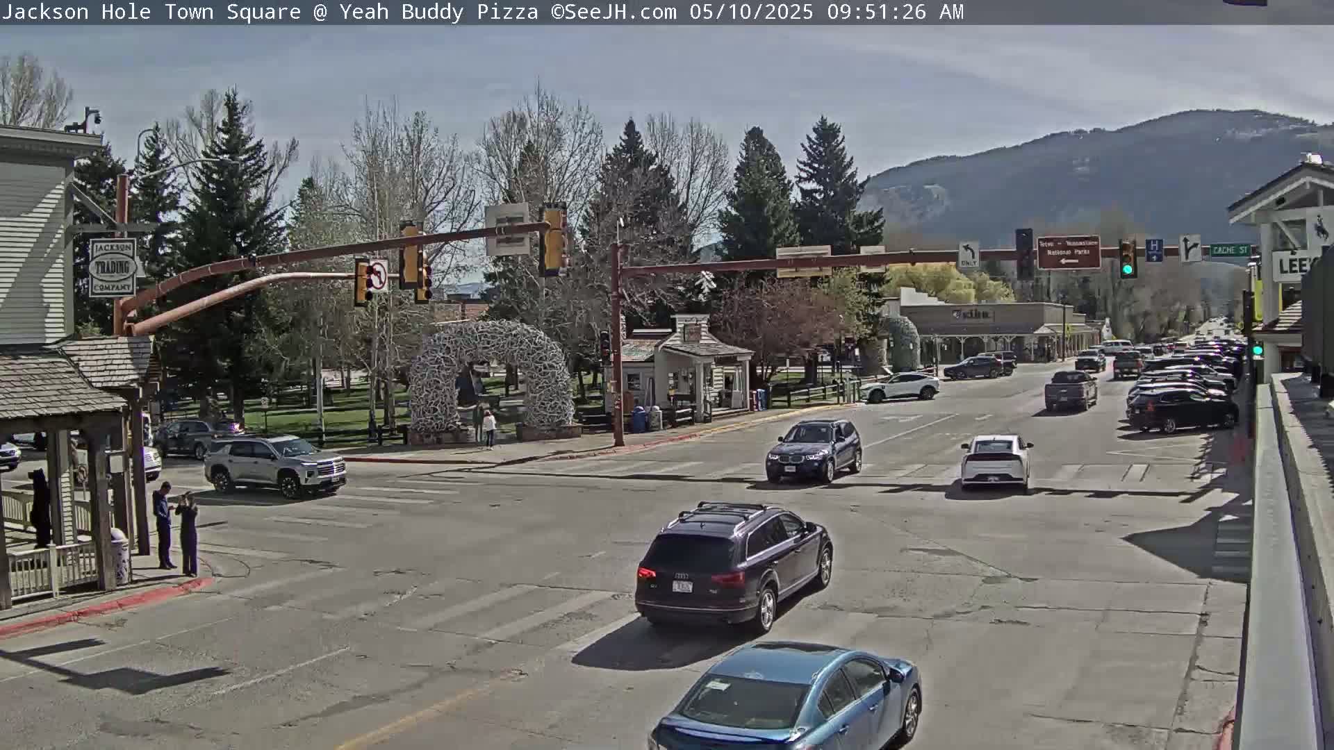 A sunny day in a town square with several cars at an intersection, surrounded by buildings and trees, with mountains in the background.