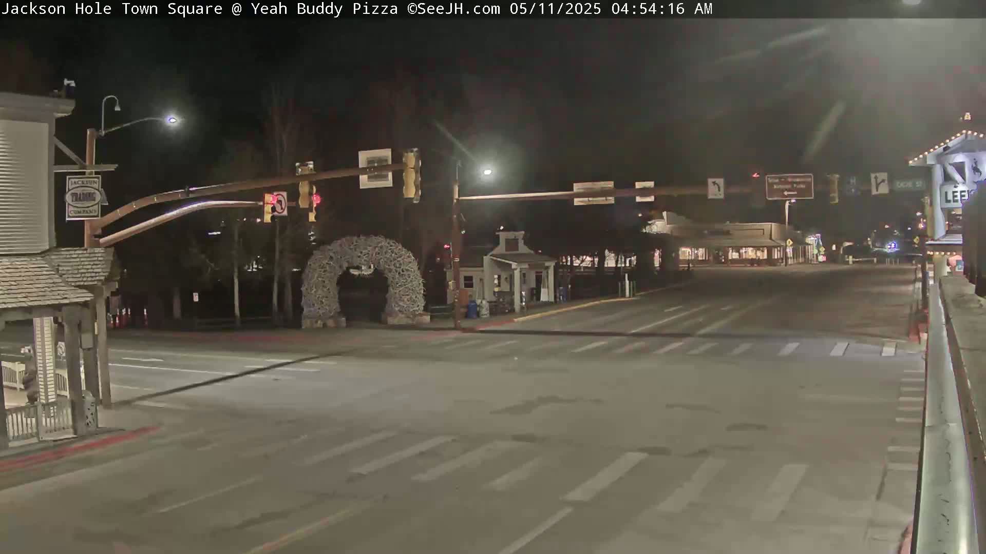 A mostly empty town square at night, illuminated by streetlights, features a large antler archway and traffic signals.