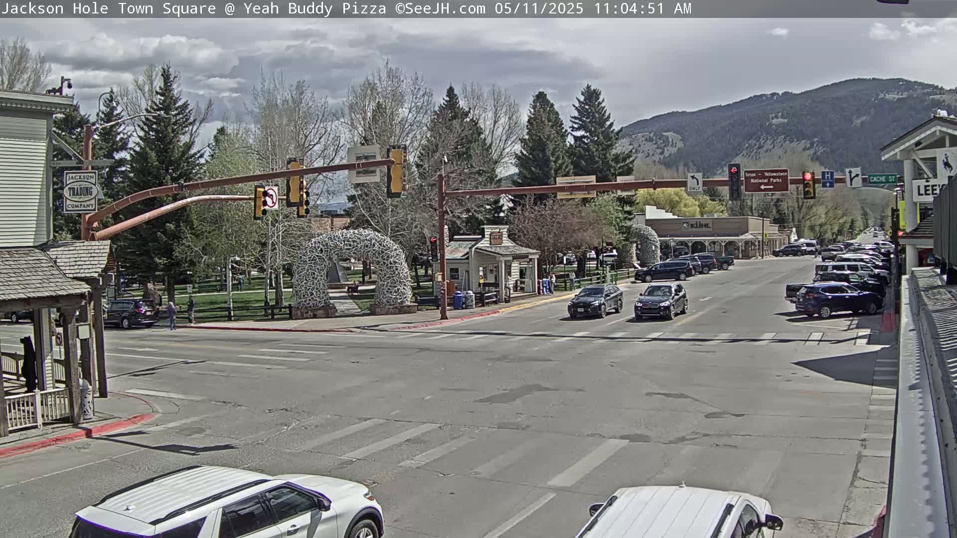 A partly cloudy day in a town square shows a street intersection with several cars, buildings, and trees, a large metal sculpture in the center, and mountains in the background.