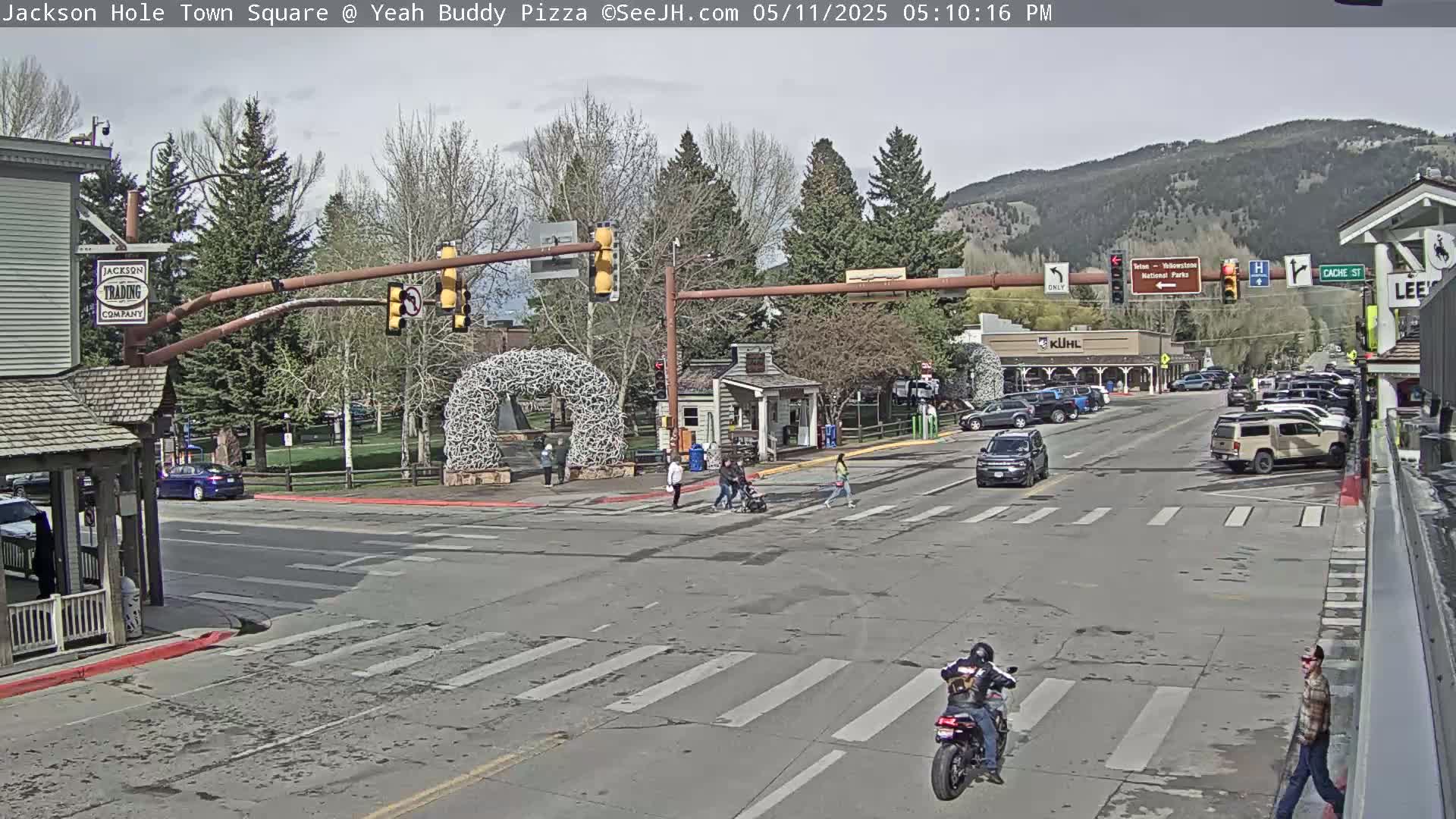 A town square intersection with pedestrians and vehicles under a partly cloudy sky, with mountains in the background.