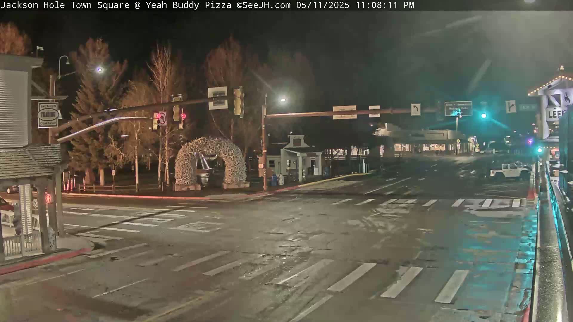 A nighttime street scene shows a mostly empty town square with traffic lights, bare trees, and a decorative arch.