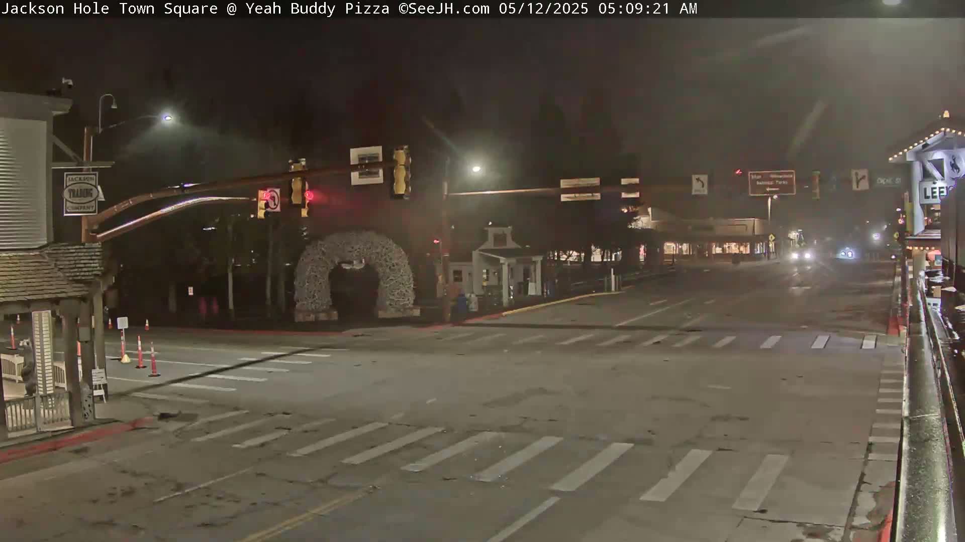 A nighttime street scene shows a mostly empty intersection with a few cars in the distance under hazy conditions.