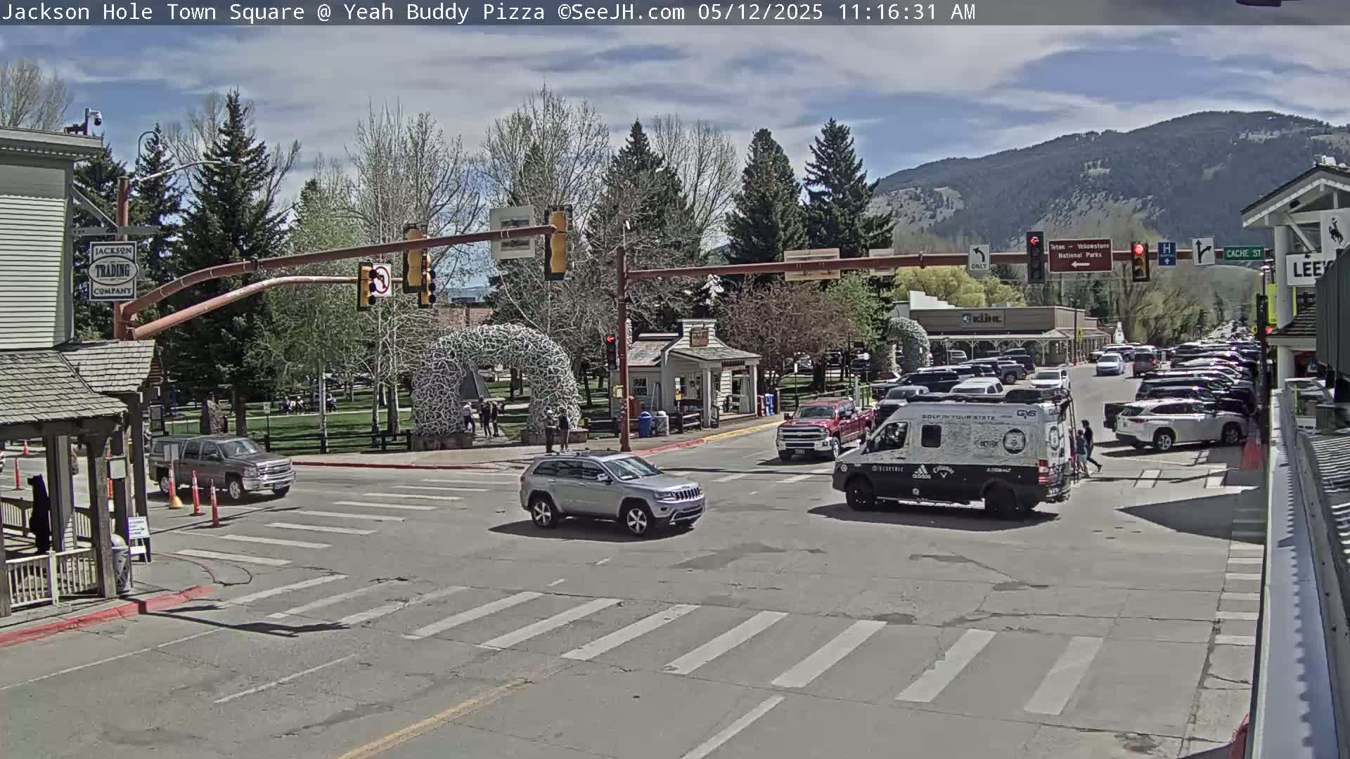 A town square intersection on a partly sunny day shows several cars at a traffic light, with parked cars lining the streets, and mountains visible in the background.