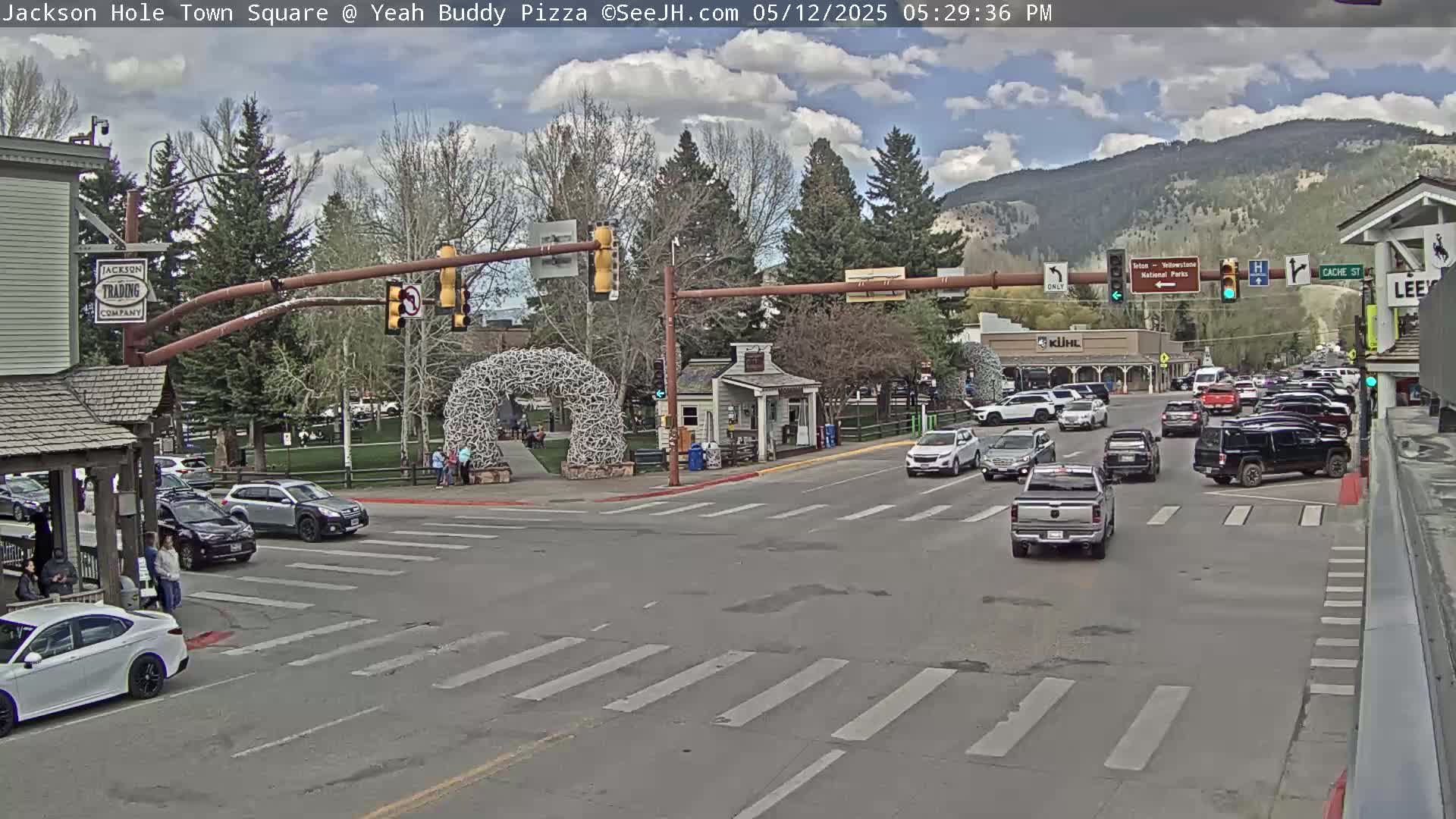A partly cloudy day in a town square shows a street intersection with several cars and pedestrians, and a large antler arch in the background with mountains in the distance.