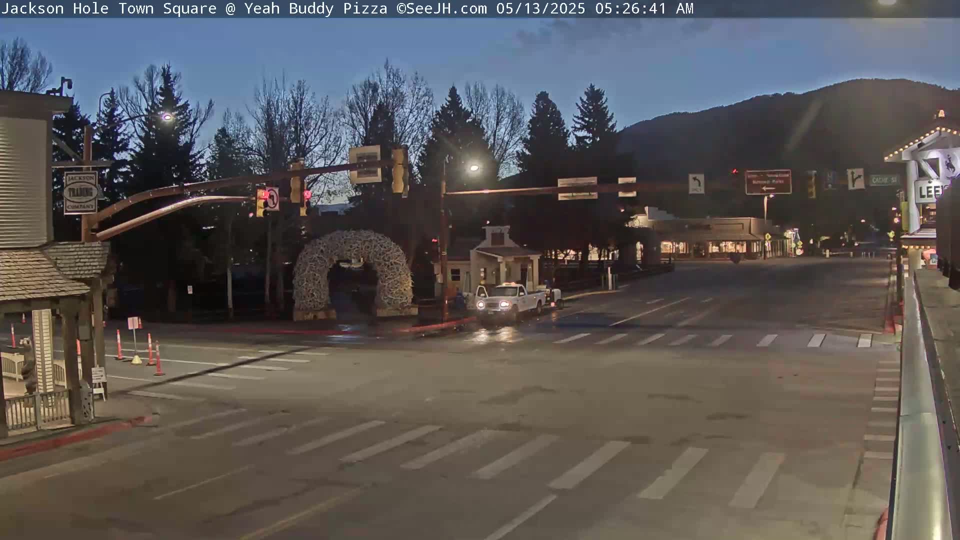 A mostly empty town square at night, under a twilight sky, with a single vehicle at an intersection near a large antler arch.