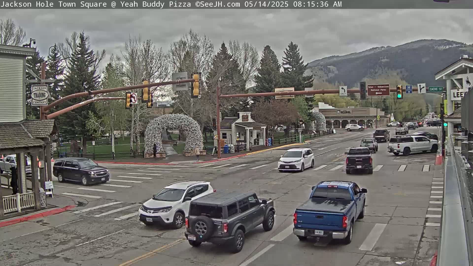 A town square intersection with several vehicles, including SUVs, pickup trucks, and minivans, under an overcast sky, with mountains in the background.
