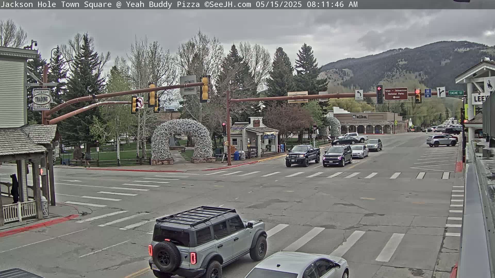 A town square intersection with several cars, under an overcast sky, features a large metal sculpture and buildings with businesses.