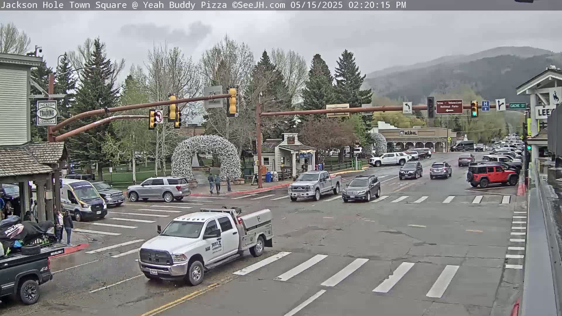 A town square intersection with several vehicles, pedestrians, and buildings under an overcast sky.