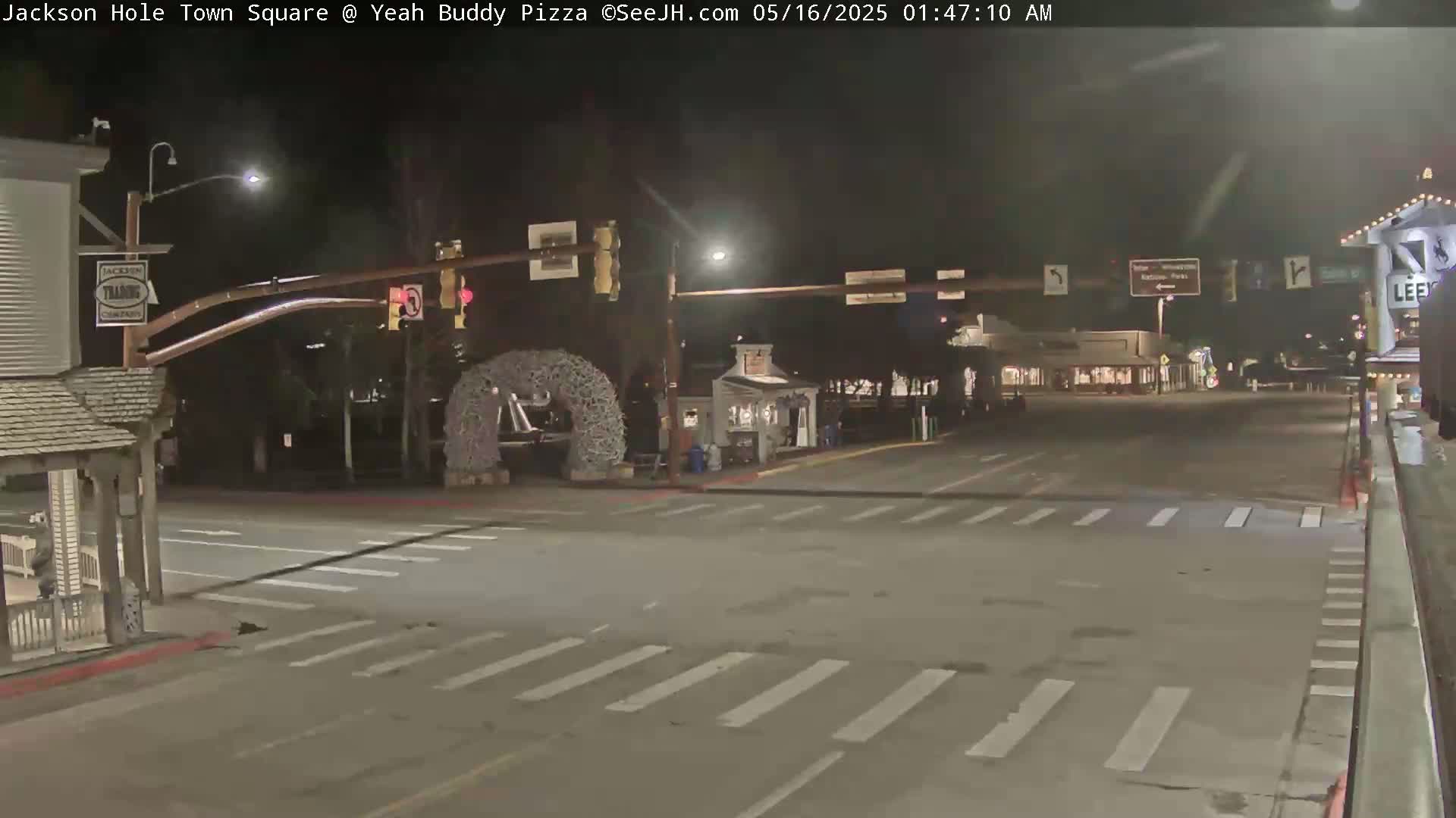 A nighttime, empty town square intersection with crosswalks and traffic signals, featuring a large decorative wreath.