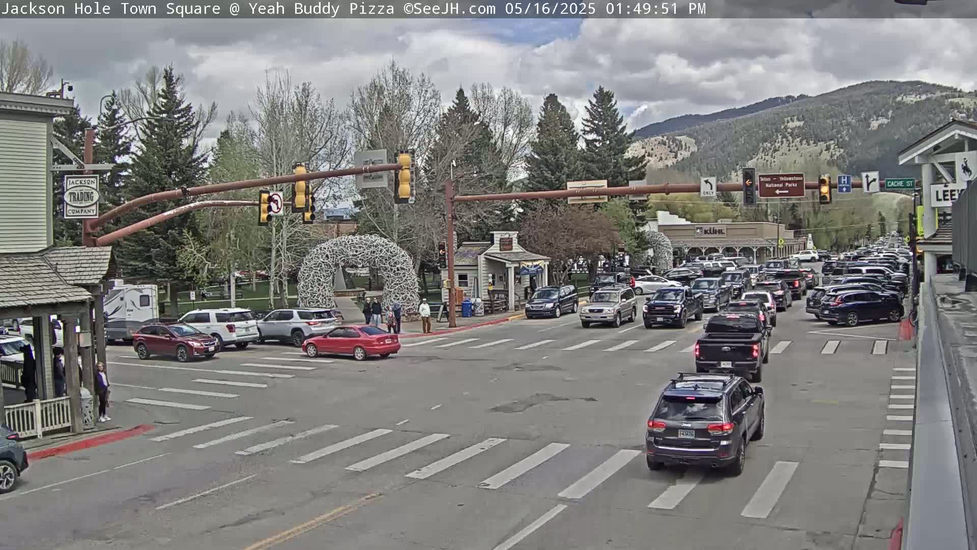 A partly cloudy day reveals a busy town square intersection with numerous vehicles, pedestrians, and buildings, including shops and restaurants, set against a backdrop of mountains.