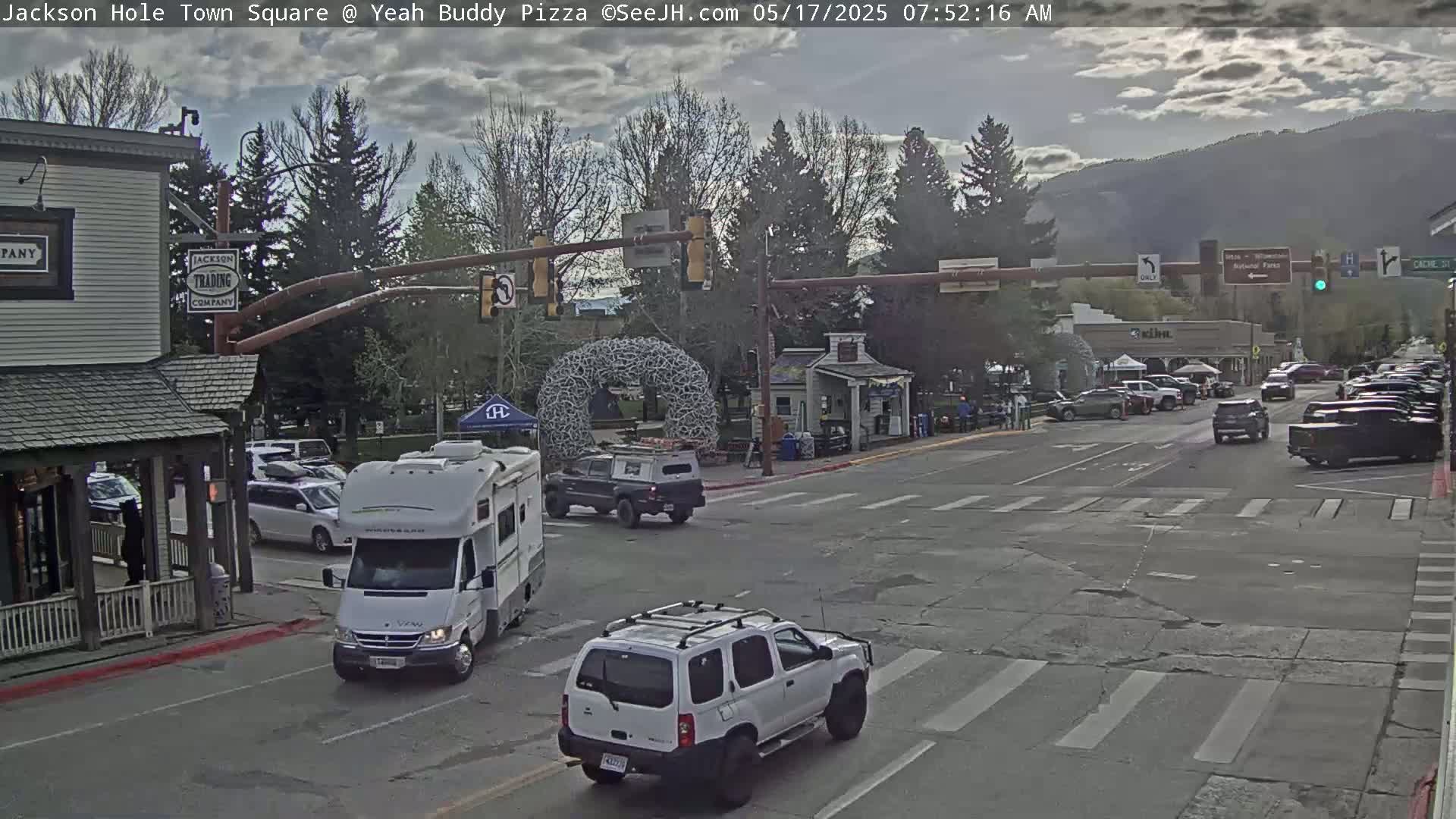 A mostly cloudy day reveals a town square with several vehicles, including an RV and SUV, at an intersection under a traffic light system; in the background are mountains and trees.
