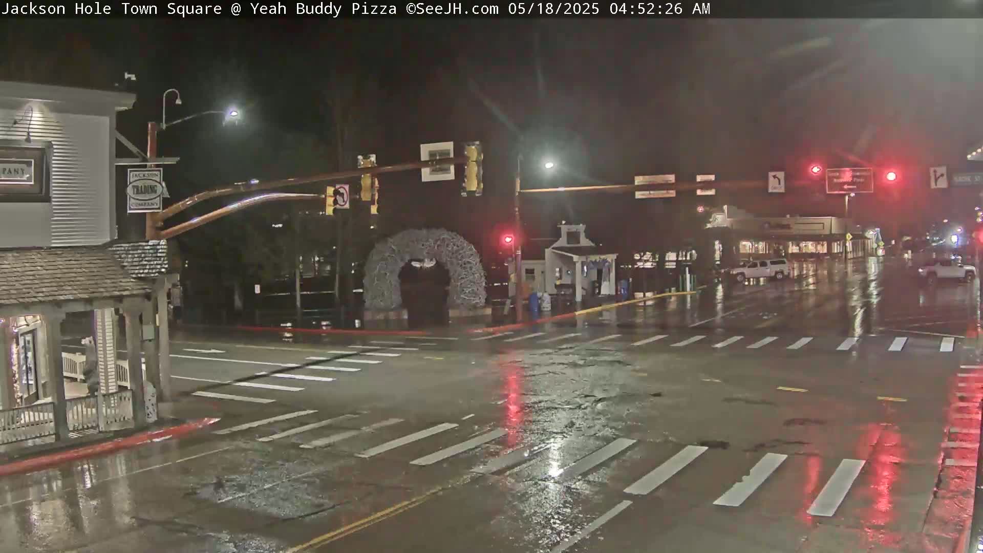 A rain-slicked town square at night, with a few vehicles visible and crosswalks marked on the wet pavement.