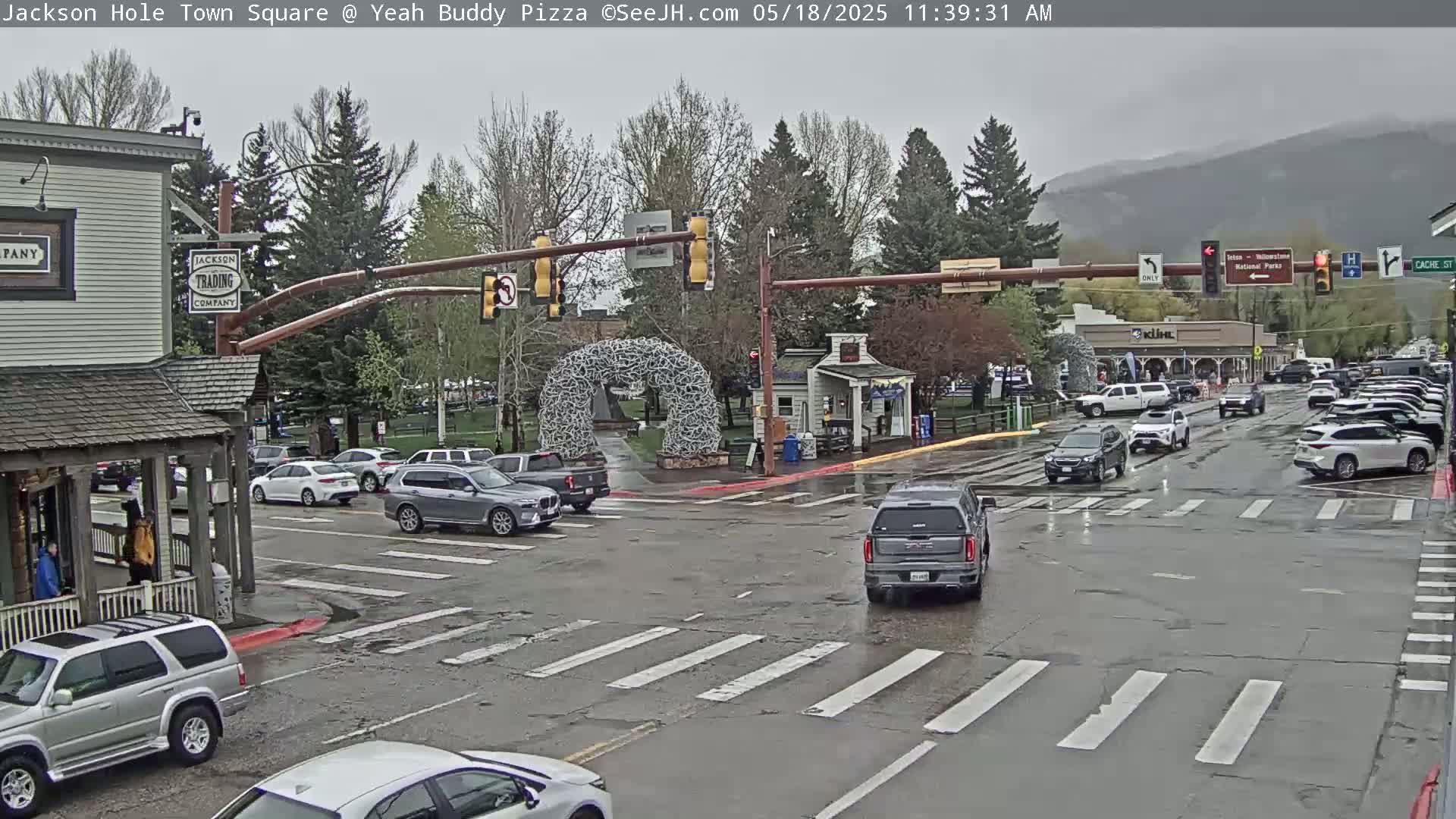 A wet town square with several cars at an intersection, under an overcast sky, features a large metal sculpture in the center.