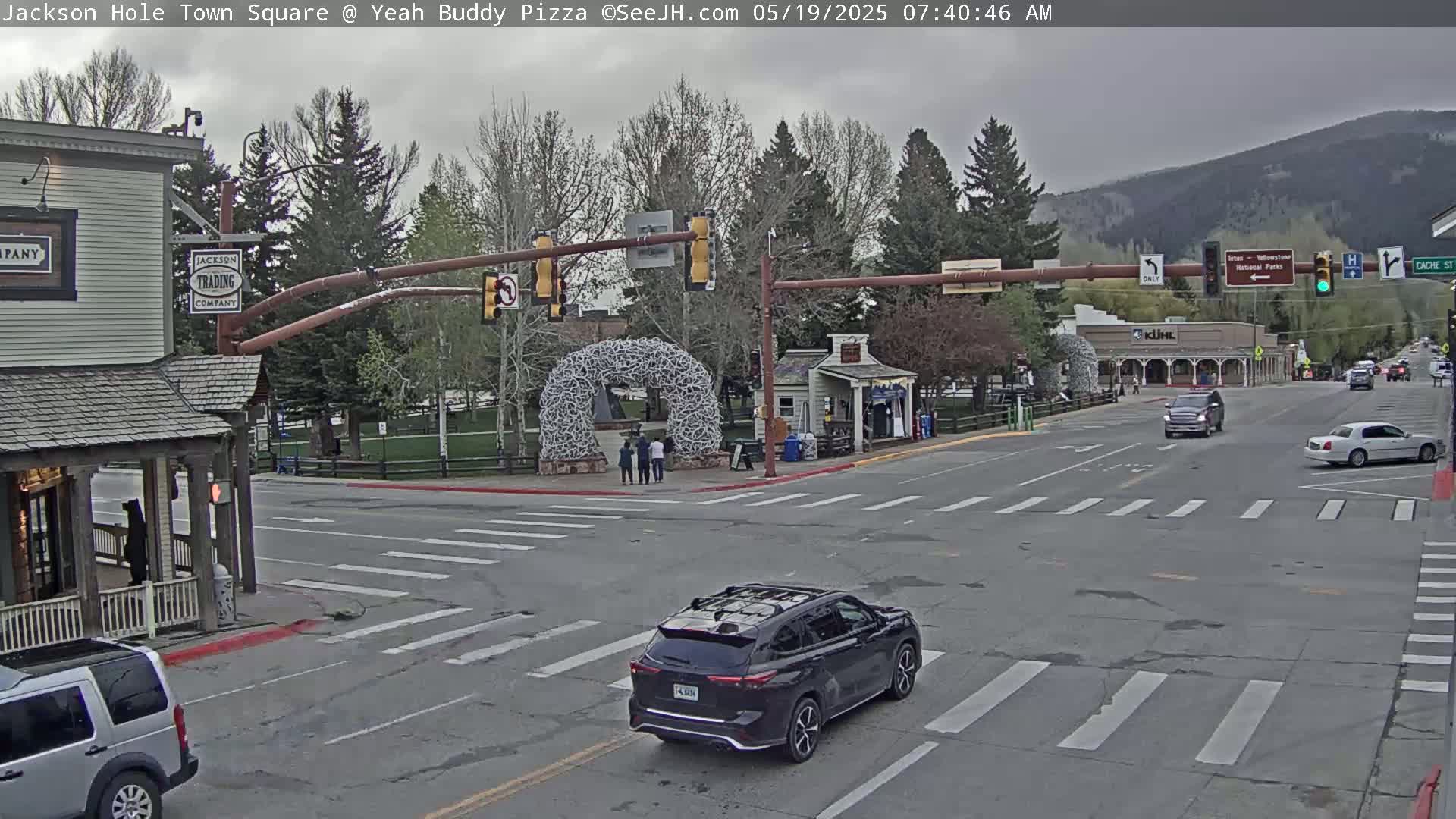 A town square intersection with overcast skies shows a dark-colored SUV stopped at a crosswalk, other vehicles traveling on the roads, and pedestrians near a large antler archway.