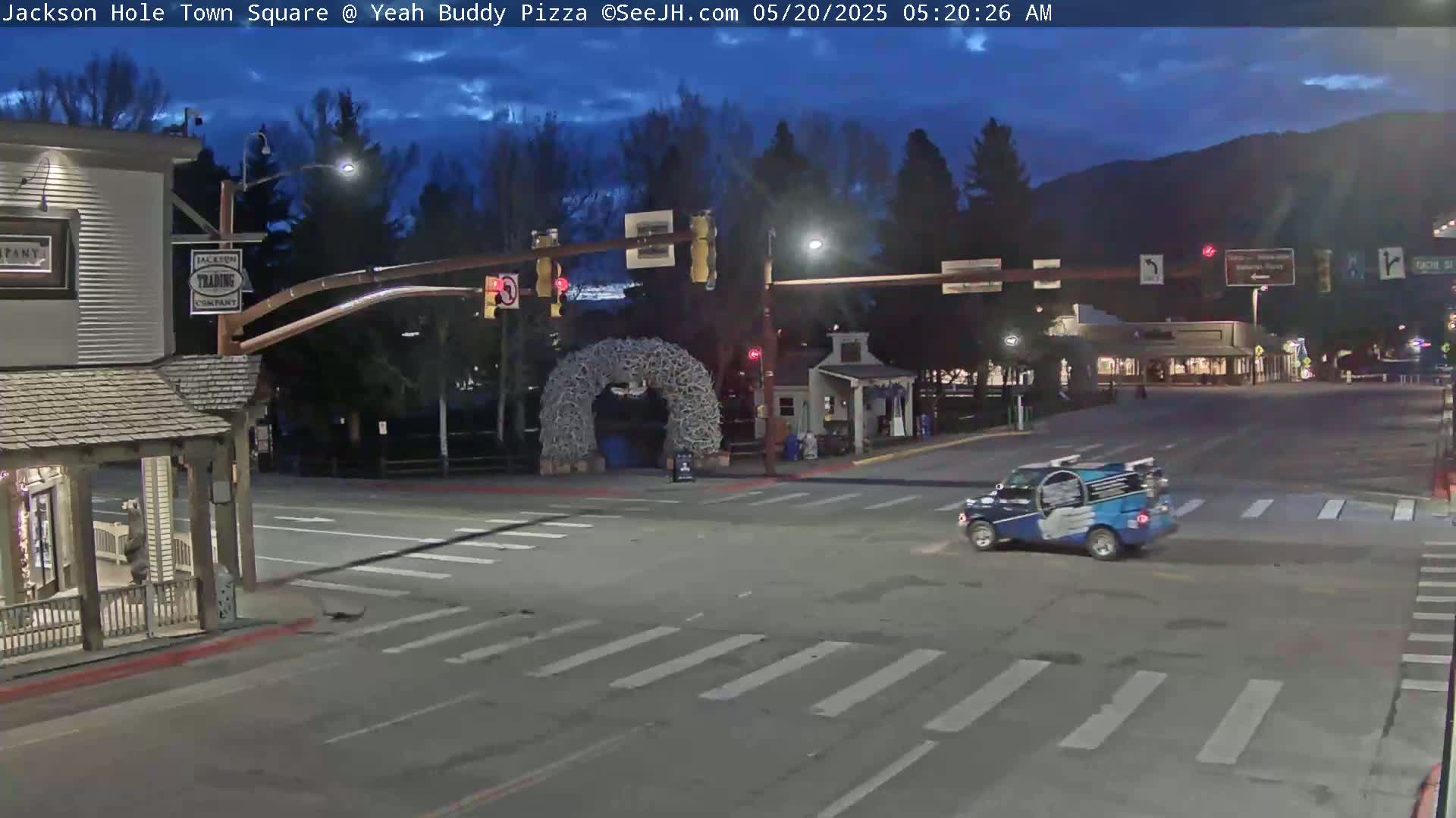A town square at dusk under a partly cloudy sky shows a vehicle driving through an intersection with crosswalks, traffic lights, and a large antler archway in the background.