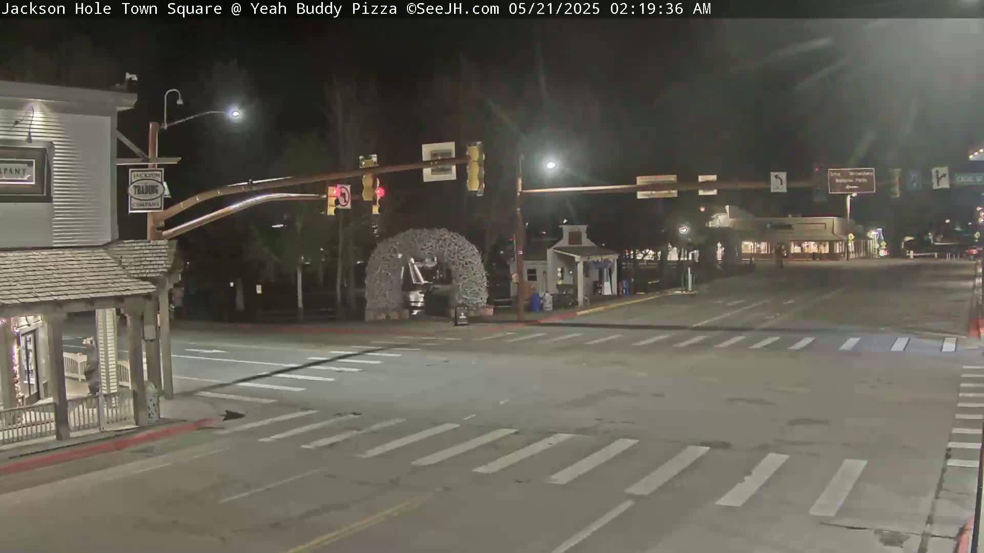 A nighttime view of a town square intersection with crosswalks, streetlights, and a large antler archway in the center.