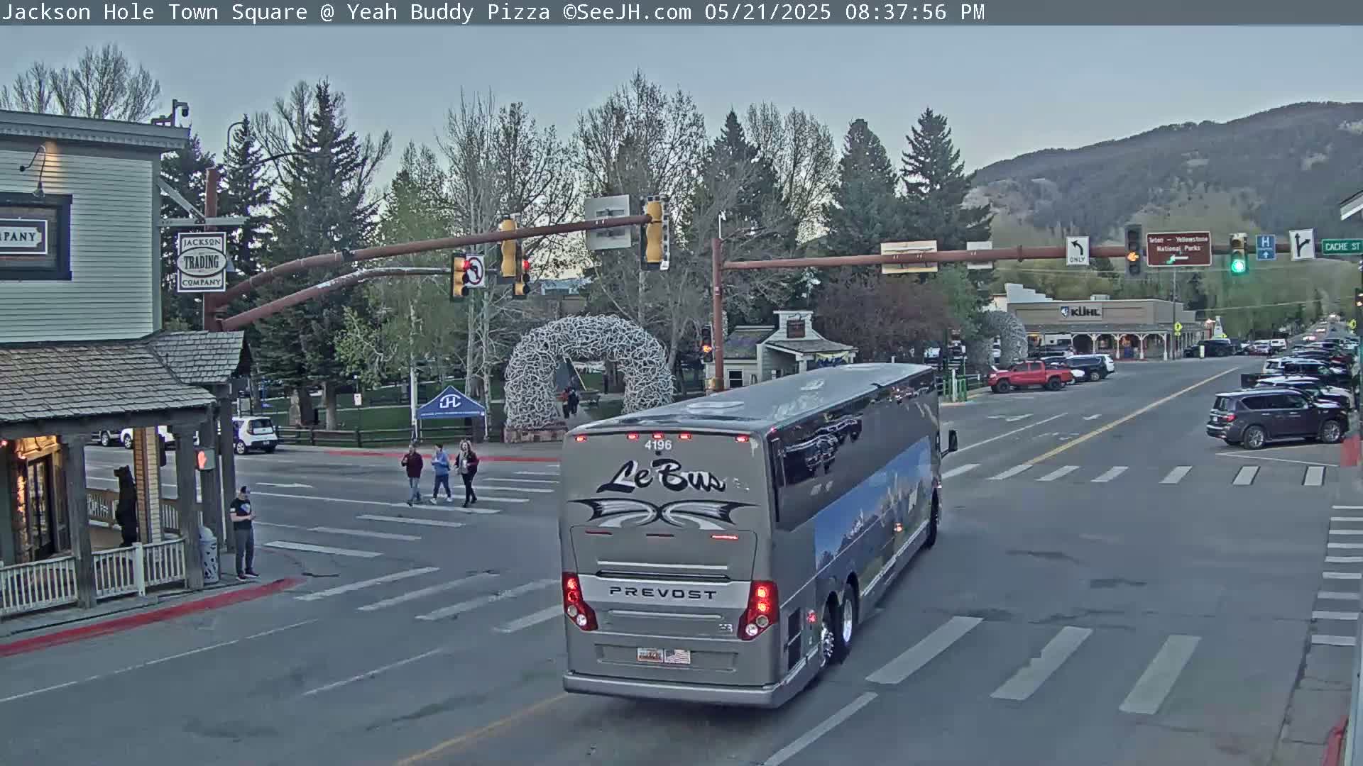 A large gray tour bus sits at a town square intersection under a partly cloudy sky, with pedestrians crossing the street and other vehicles nearby.