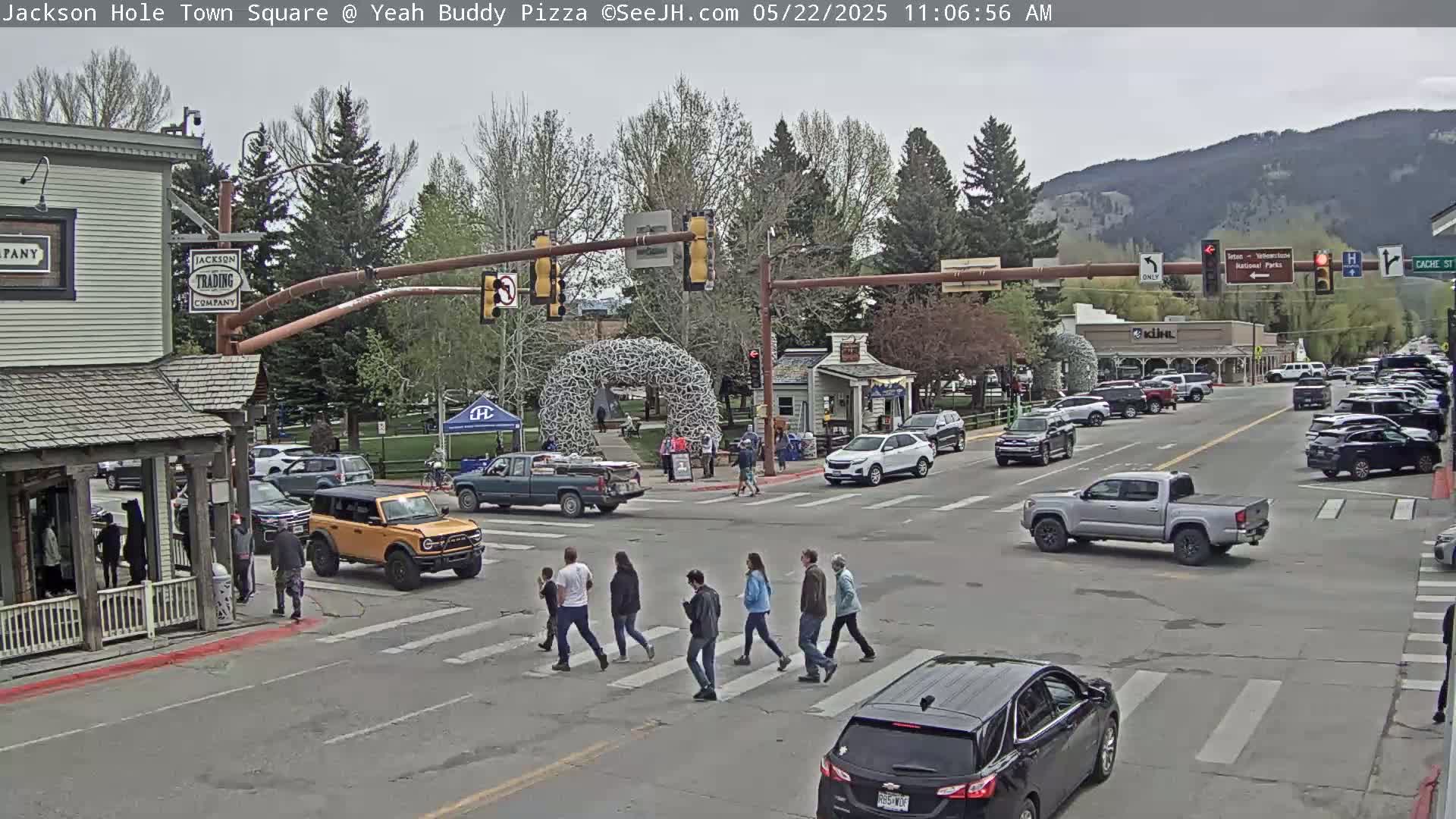 A town square intersection with several vehicles and pedestrians, under an overcast sky, with mountains in the background.