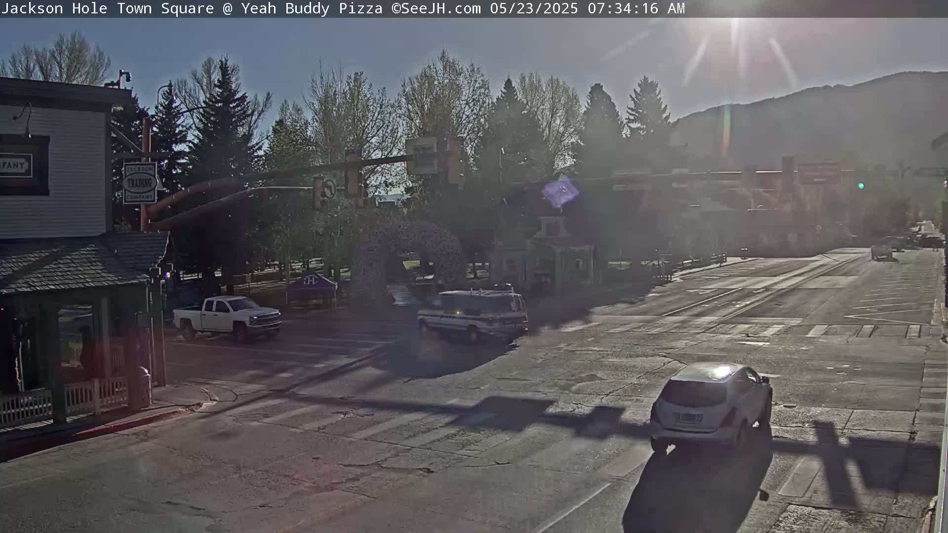 A sunny morning view of a town square intersection with several vehicles, including a white pickup truck and a white SUV, amidst buildings and trees.