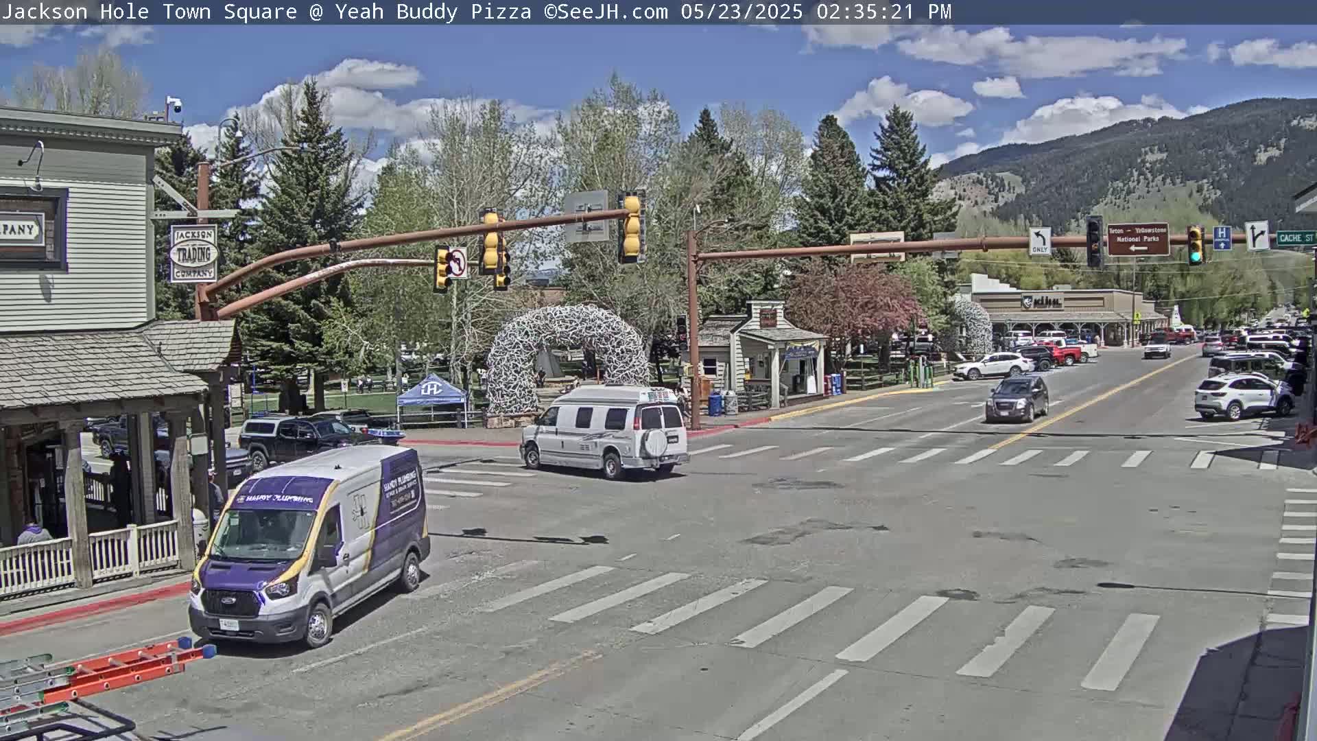 A partly sunny day shows a town square intersection with several vehicles, including vans and cars, with a large antler arch in the background and mountains in the distance.