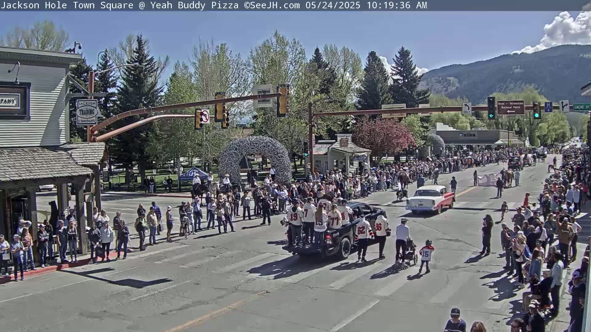 A large crowd lines the streets of a town square on a sunny day, watching a parade featuring a vehicle carrying people in hockey jerseys.