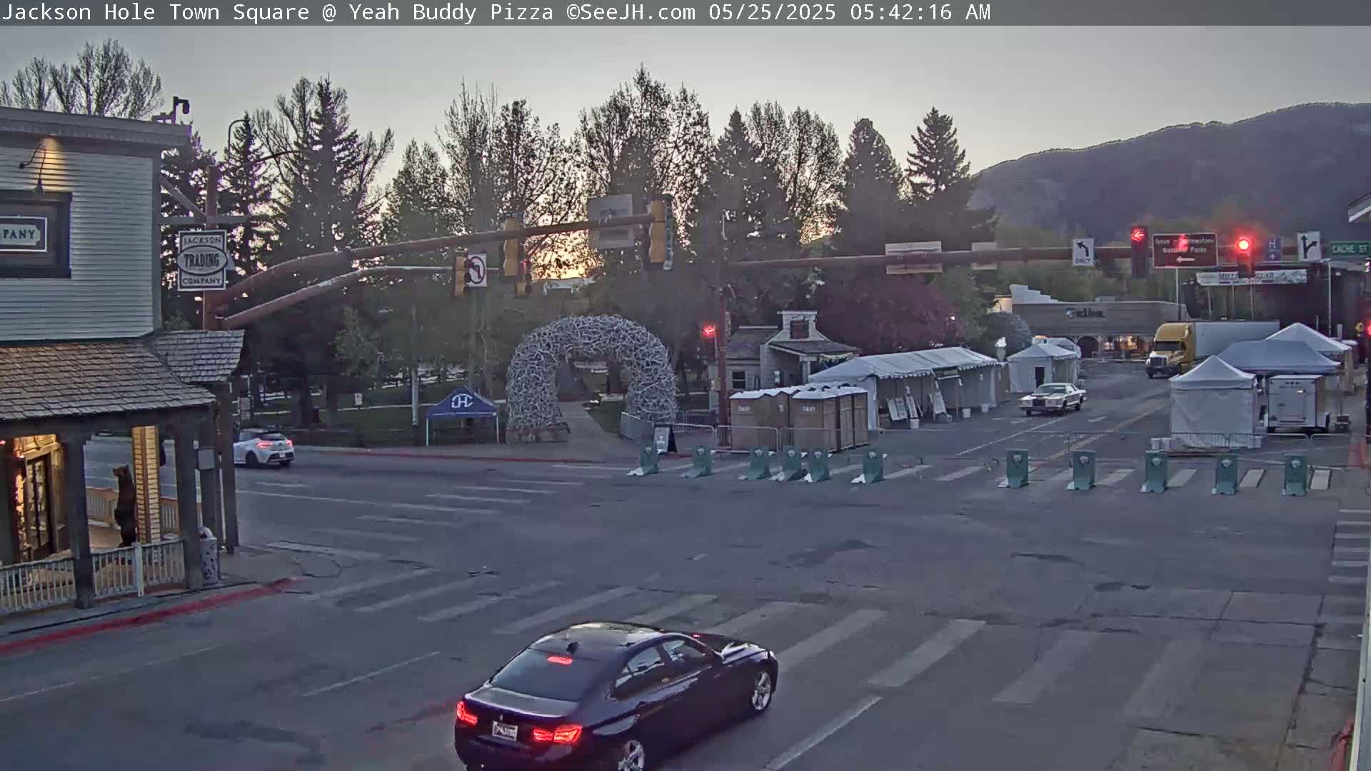A town square at sunrise shows several cars, a large antler arch, temporary vendor tents, and buildings, under a mostly clear sky.