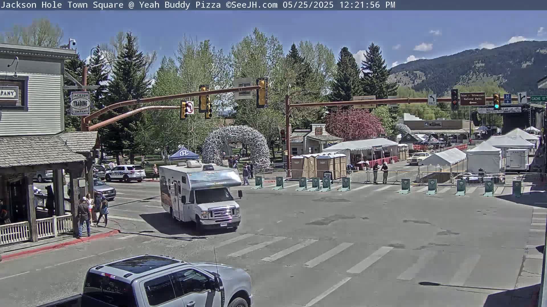 A sunny town square scene shows a street intersection with several vehicles, pedestrians, and temporary vendor tents set up against a backdrop of mountains and trees.