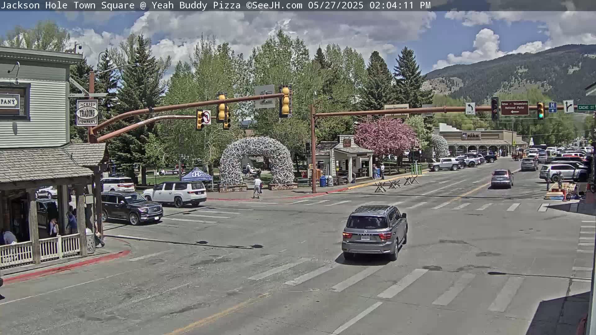 A partly cloudy day in a town square shows cars at an intersection, with buildings, trees, and a large antler arch visible.