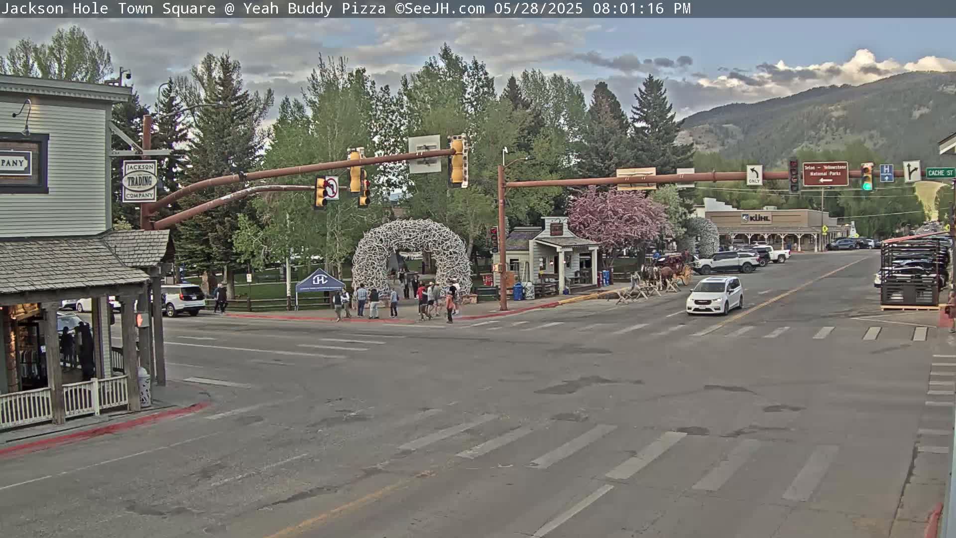 A town square with a few pedestrians and vehicles under partly cloudy skies, features a large antler archway and buildings lining the street.