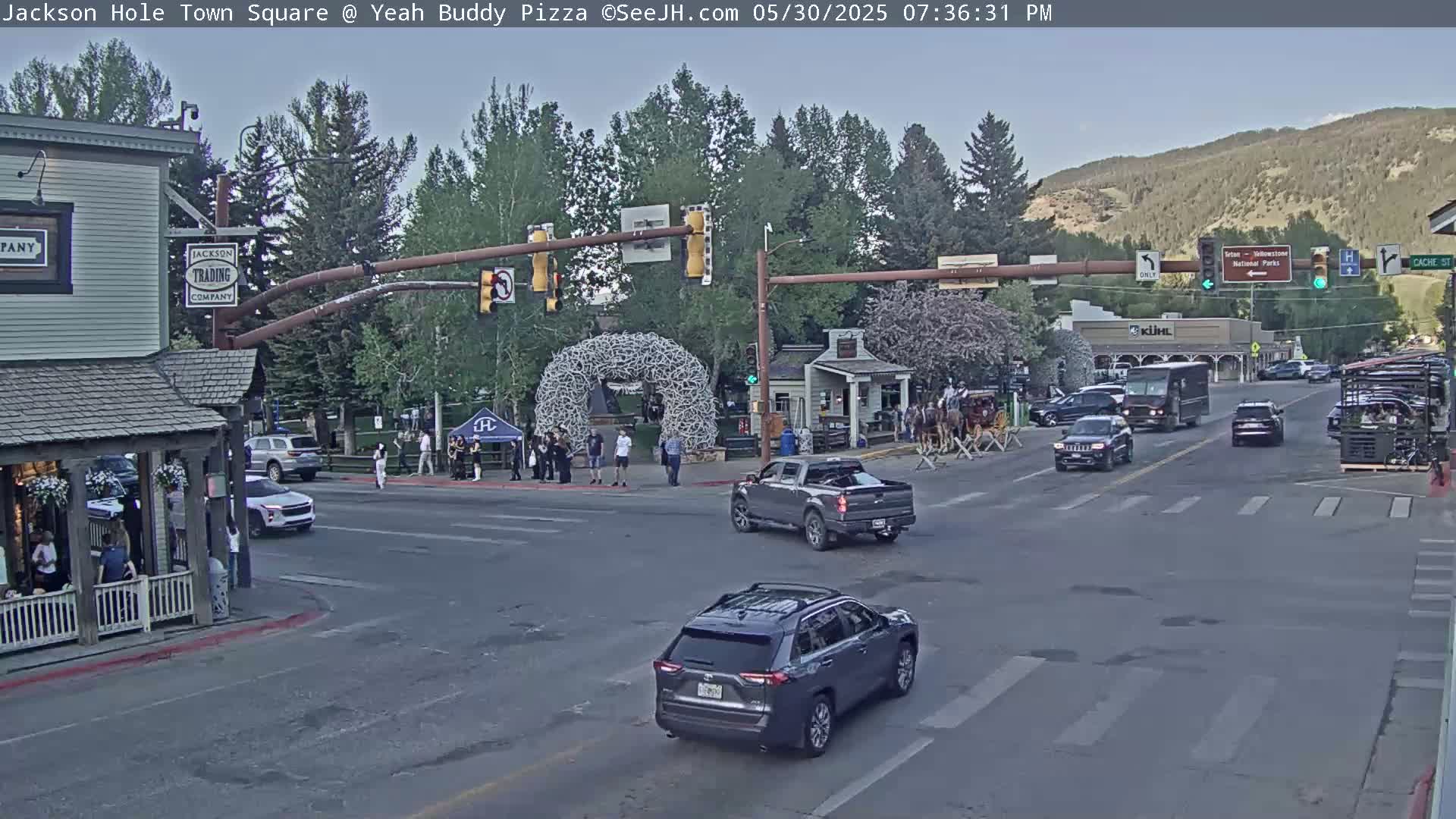 A town square intersection with several vehicles, pedestrians, and a large antler archway, under a partly cloudy evening sky.