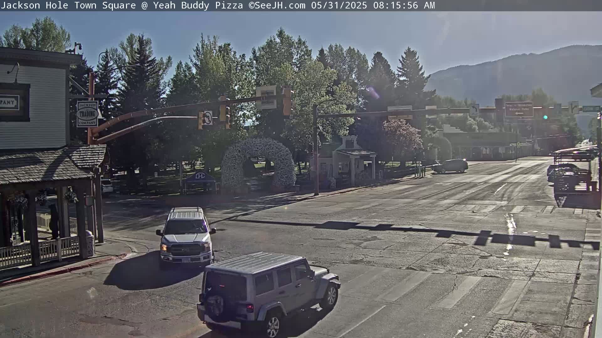 A sunny morning scene shows a town square intersection with several vehicles, including a pickup truck and a Jeep, amidst buildings and trees, with mountains in the background.