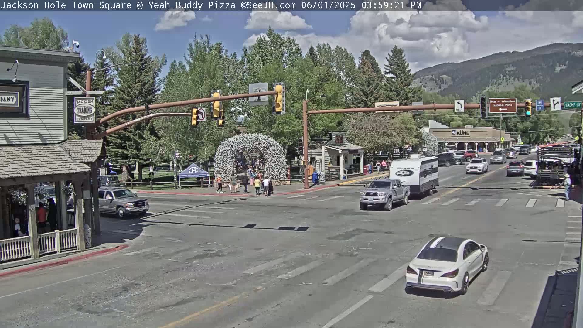 A town square intersection with several vehicles and pedestrians under a partly cloudy sky.