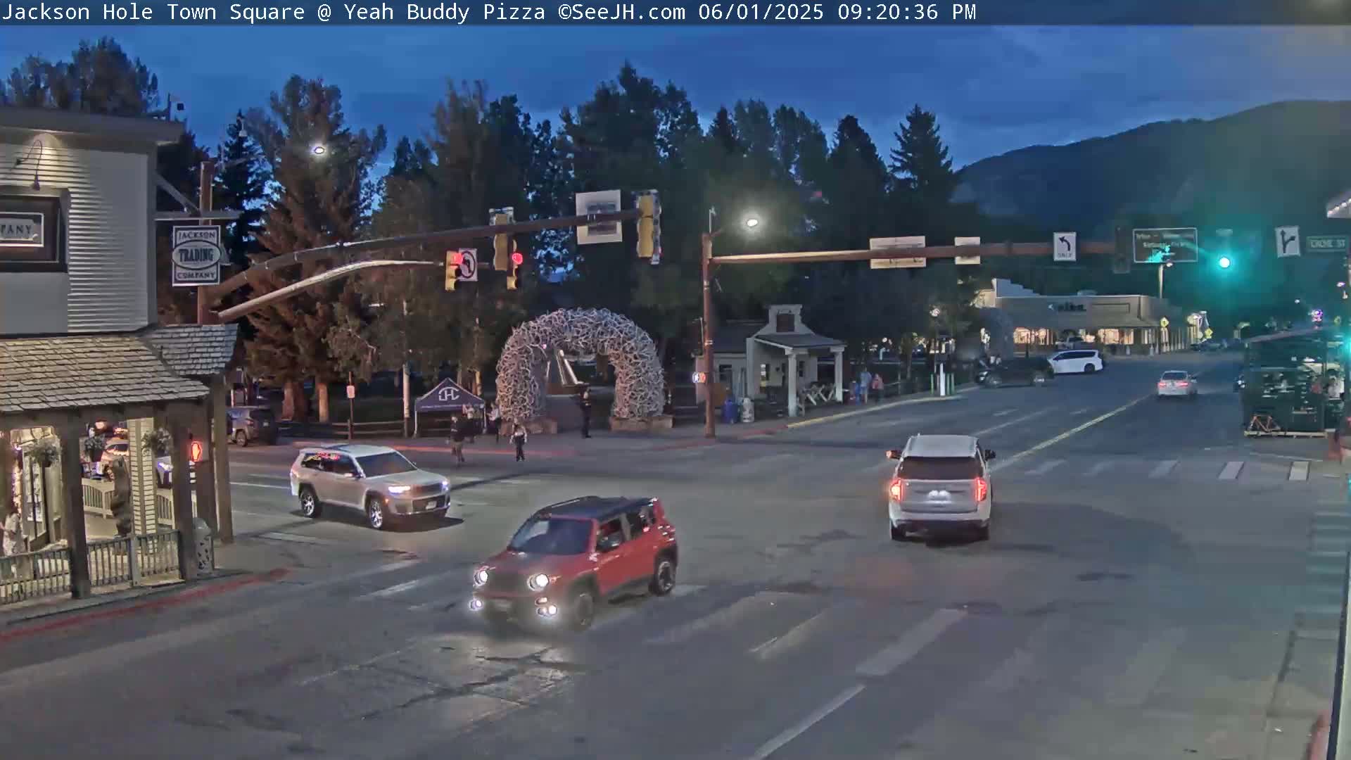 A town square at dusk with several vehicles at an intersection, a large antler archway in the center, and mountains visible in the background under a twilight sky.
