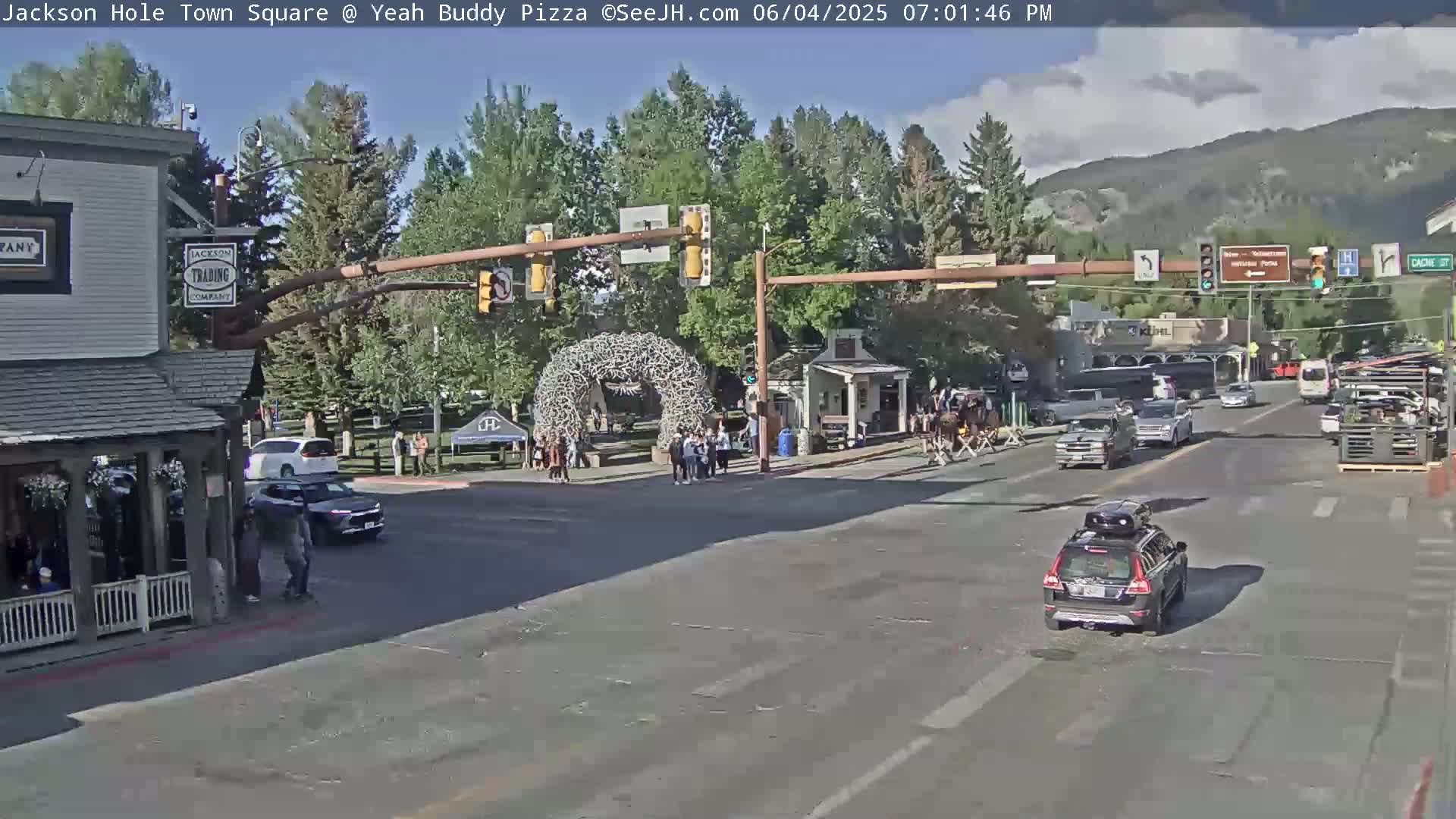 A sunny town square scene shows a street intersection with pedestrians, cars, and buildings, including one with a large antler archway.