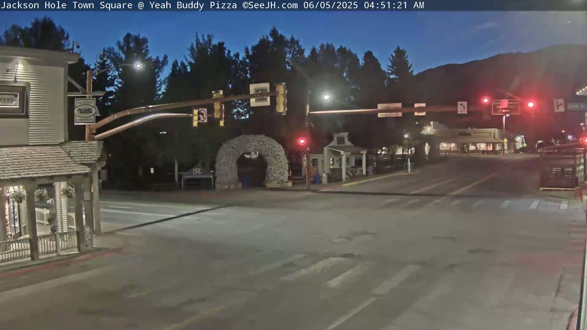 A nighttime view of a town square intersection with traffic lights, buildings, and a large antler archway under a twilight sky.