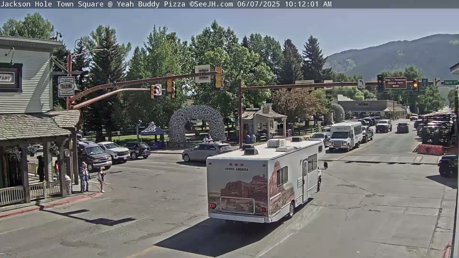 A sunny day in a town square shows a motorhome and other vehicles at an intersection, with buildings and trees surrounding a park-like area.