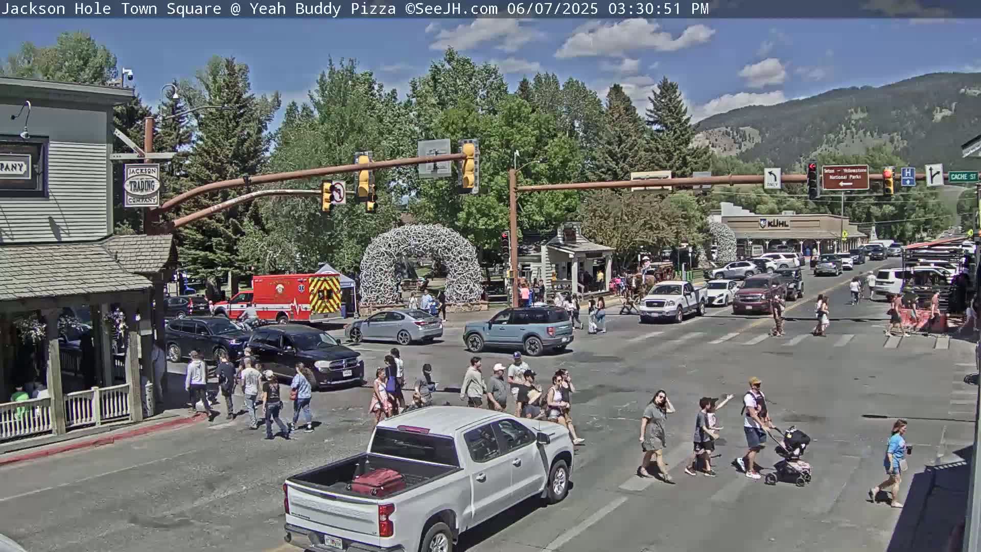 A sunny town square scene shows a busy intersection with pedestrians crossing, cars driving, and a firetruck parked near the side of the road.