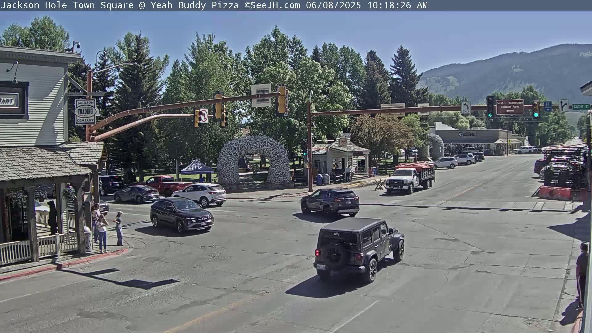 A sunny town square with several cars at an intersection, surrounded by buildings, trees, and mountains in the background.