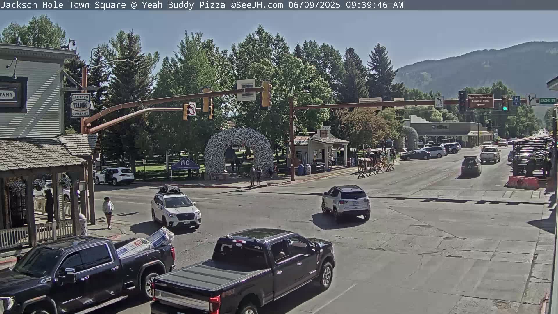 A sunny day in a town square shows several vehicles at an intersection, with pedestrians walking near buildings and trees, and mountains in the background.