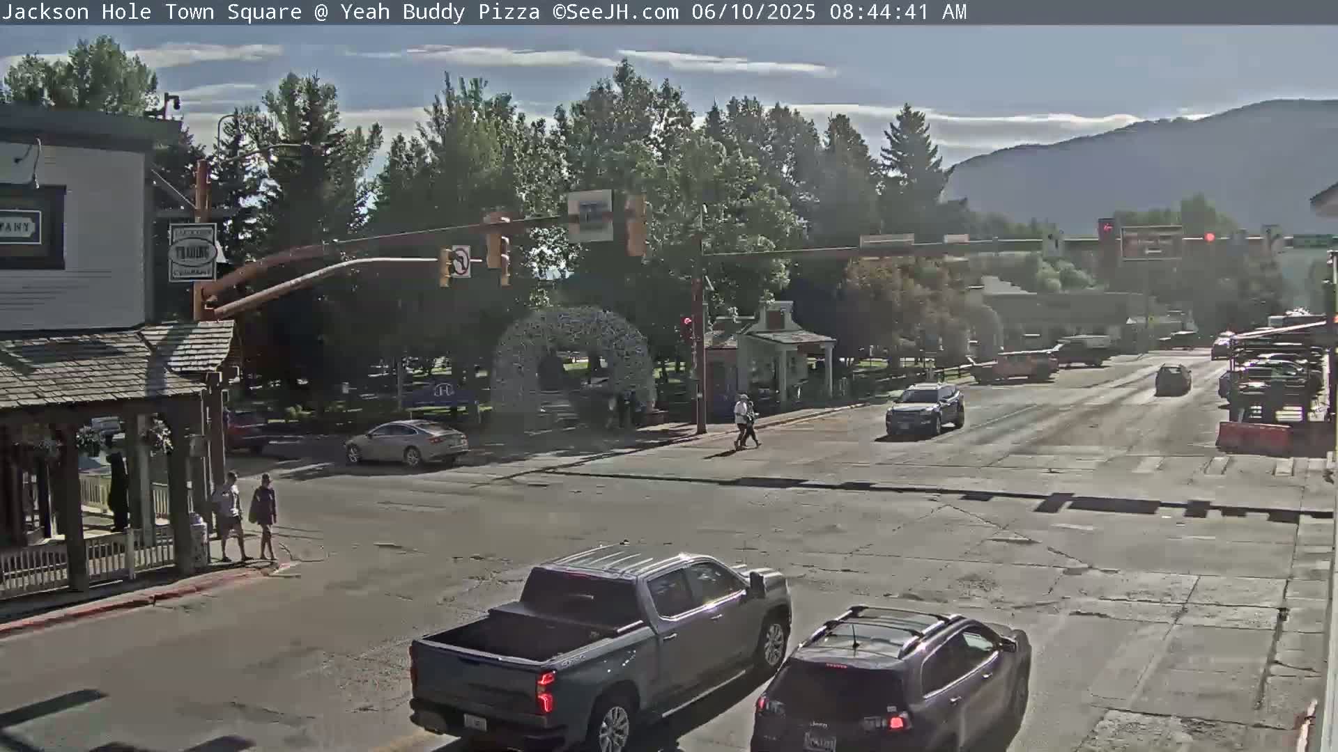 A sunny day in a town square shows a few pedestrians crossing the street and several vehicles at an intersection, with mountains visible in the background.